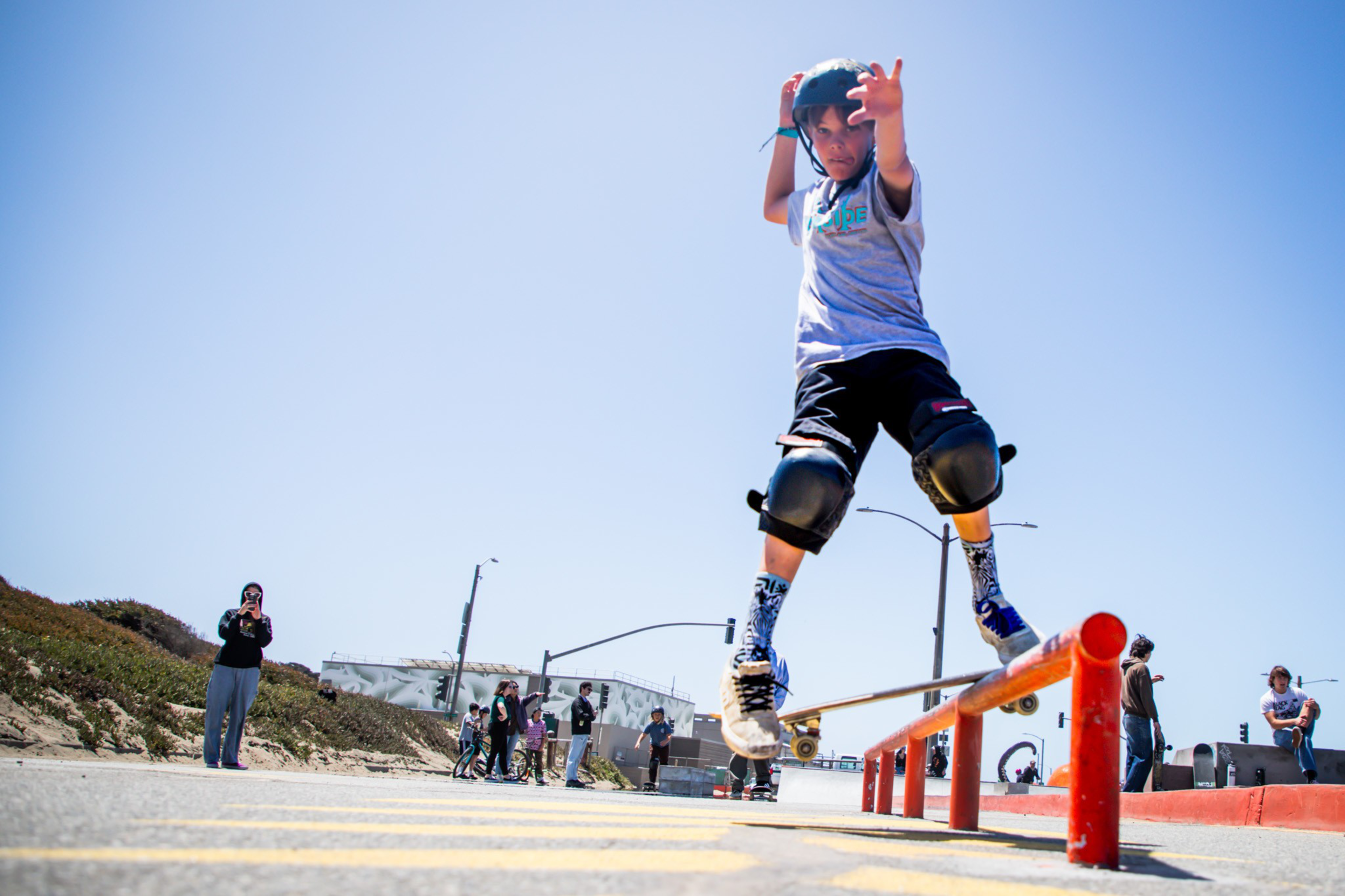A young skateboarder, wearing a helmet and knee pads, performs a trick on a red rail under a clear blue sky, with onlookers in the background.
