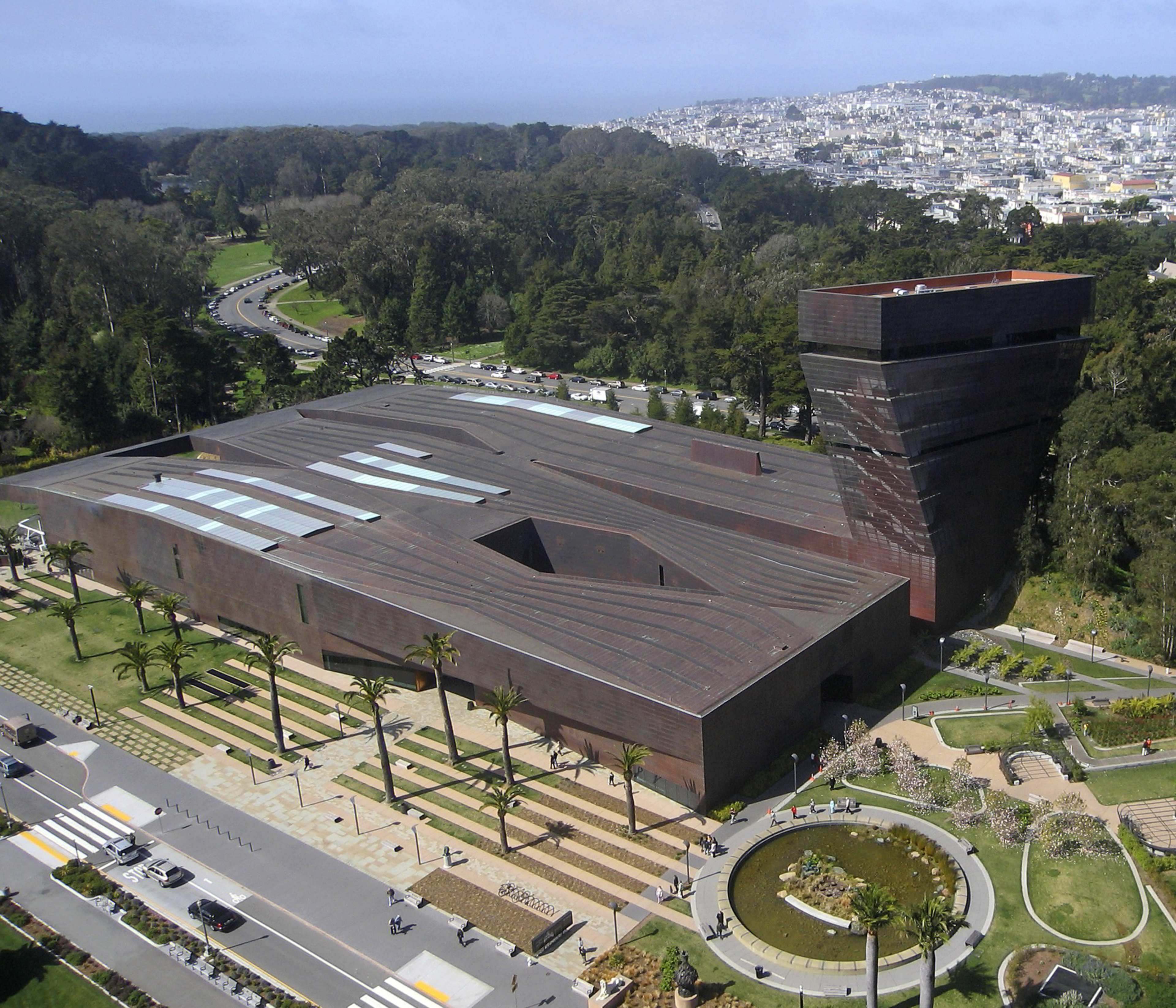 The image shows a modern, angular building with a tall tower, set in a landscaped area with trees and paths. Nearby, there’s a circular pond and cityscape in the distance.