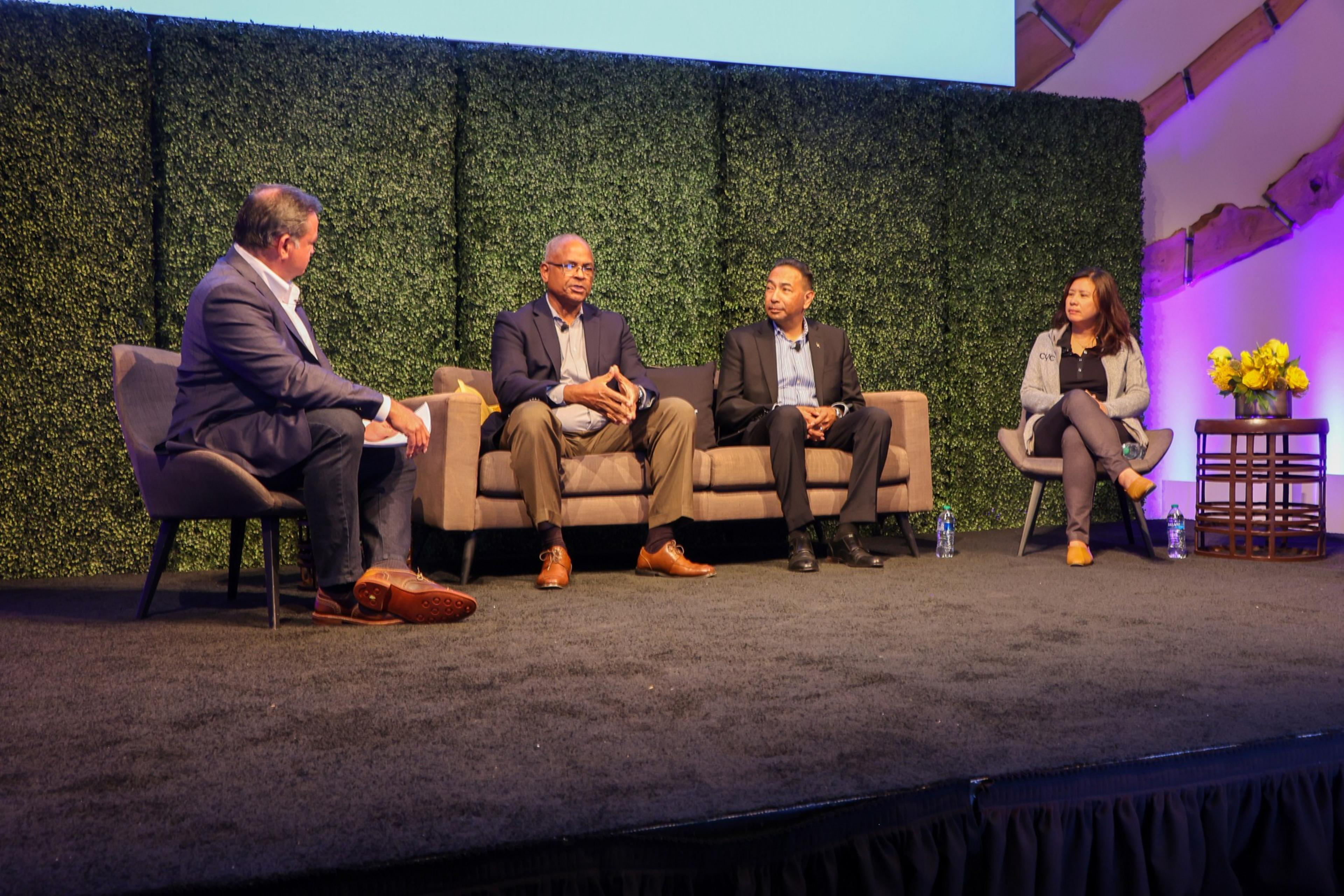 Four people are on stage, seated for a discussion, with a greenery backdrop and soft lighting.