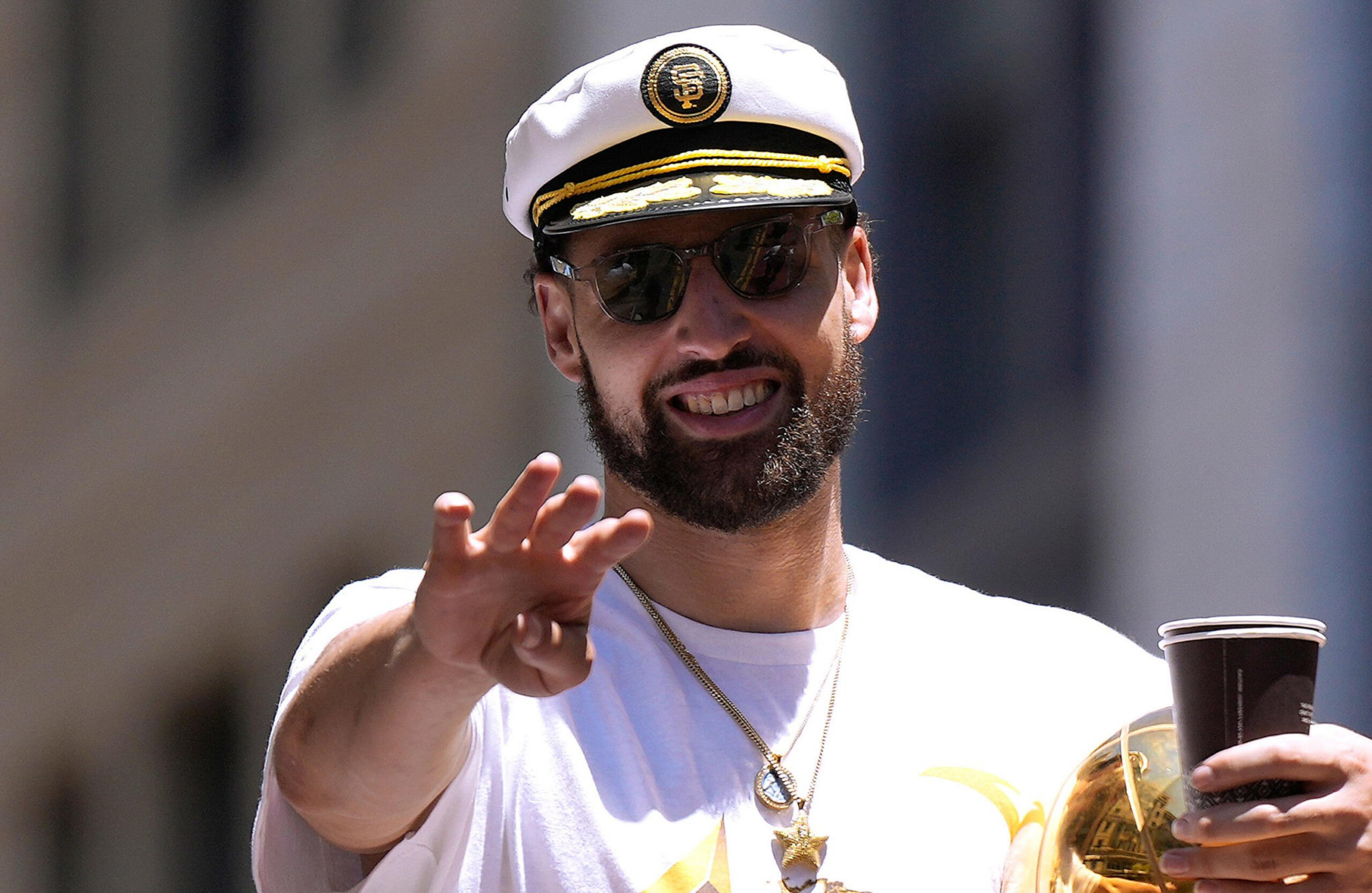 A bearded man wearing a white captain's hat and sunglasses smiles while holding a drink cup and a shiny brass trophy, reaching out with his hand.