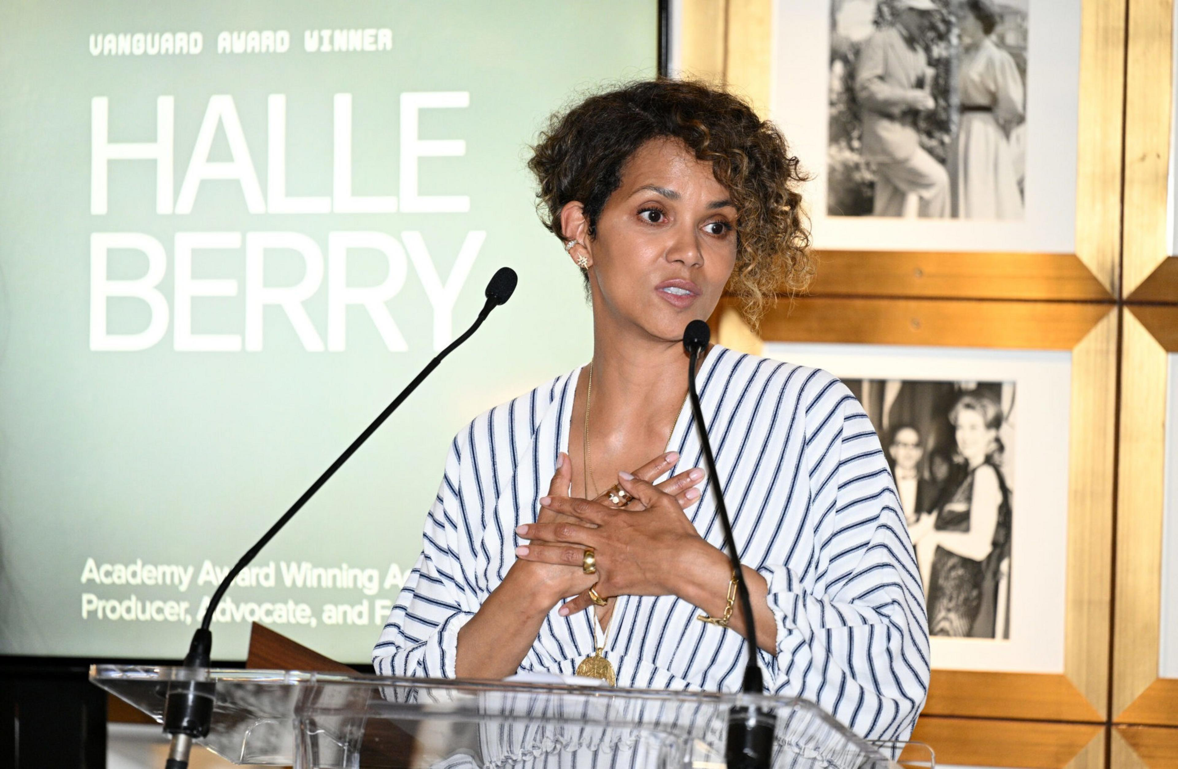 Halle Berry stands at a podium, speaking into a microphone with hands clasped, wearing a striped white and blue outfit with framed photos behind her.