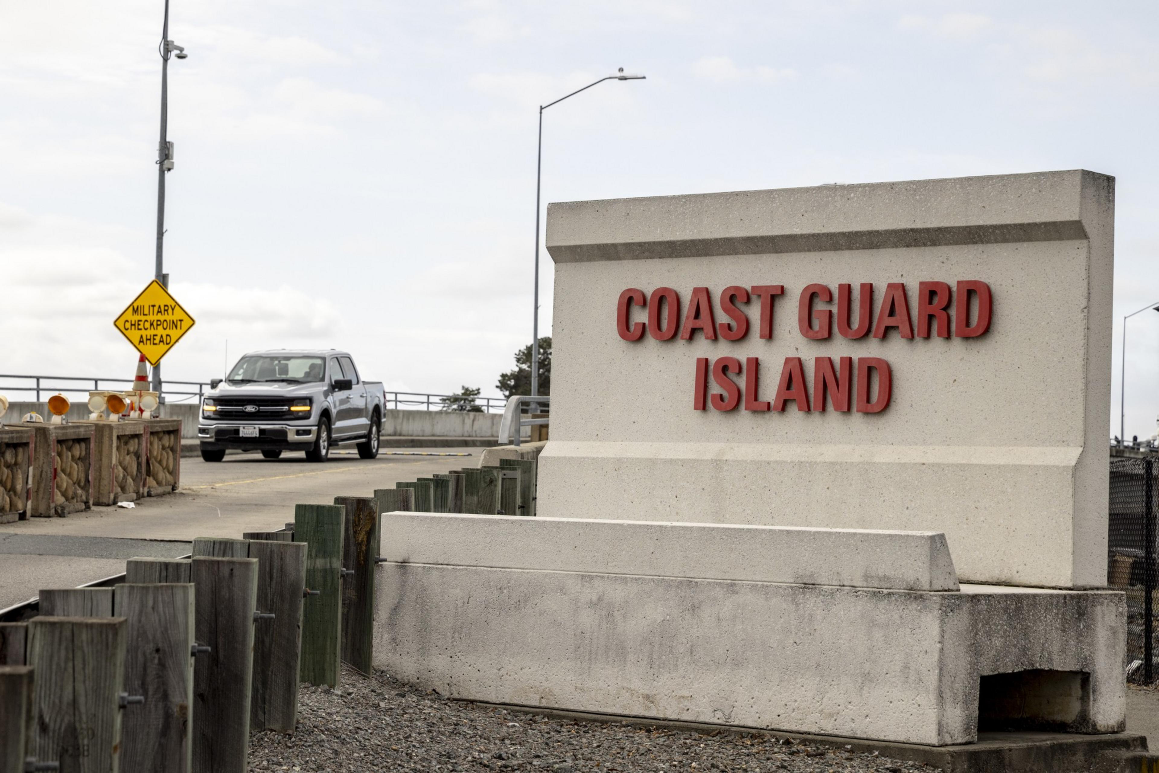 A concrete sign with red letters reads "COAST GUARD ISLAND" by a road where a gray pickup truck is driving past a yellow "MILITARY CHECKPOINT AHEAD" sign.