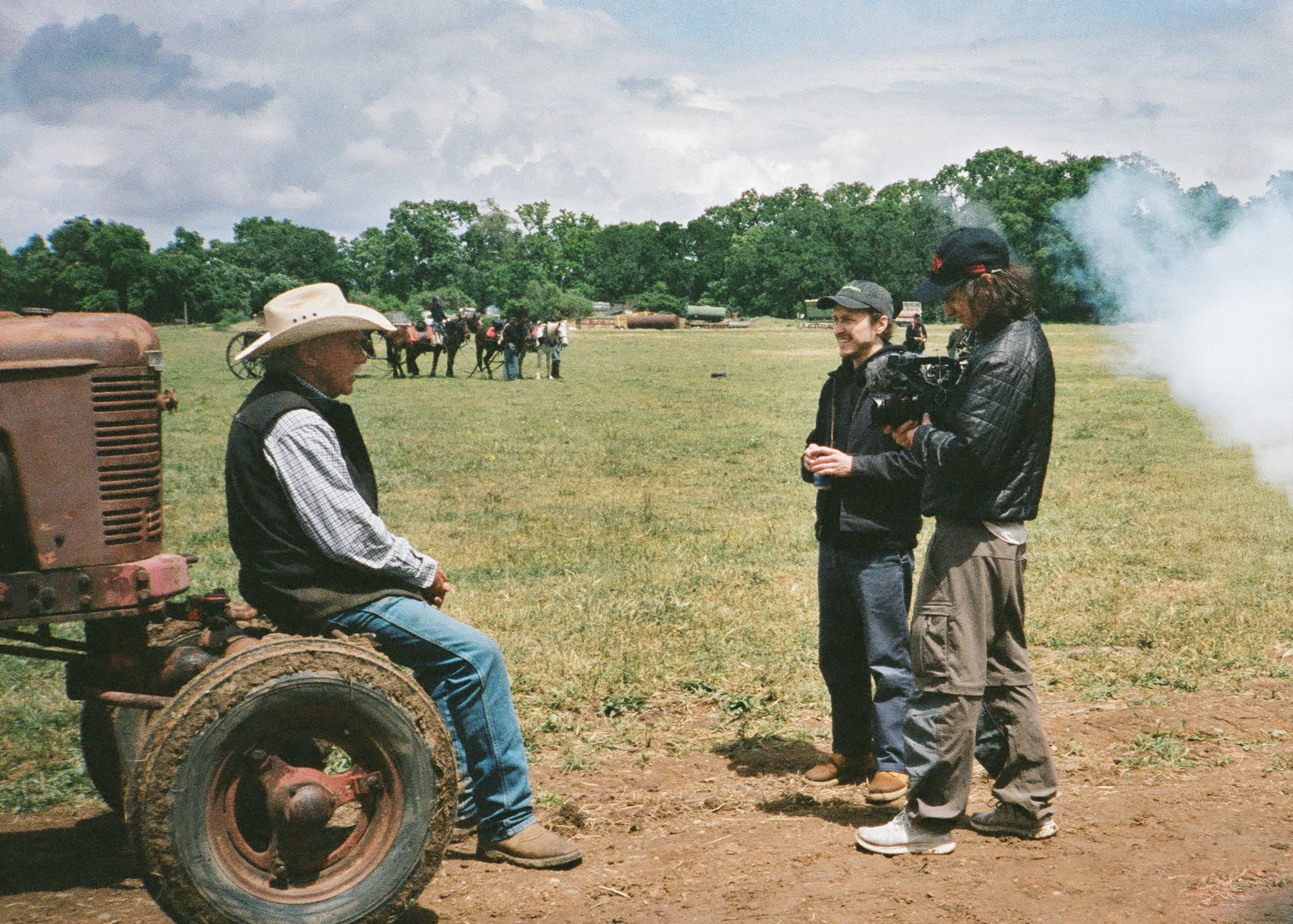 A man in a cowboy hat sits on a tractor talking to two people, one with a camera. In the background, there are horses and people on a grassy field.