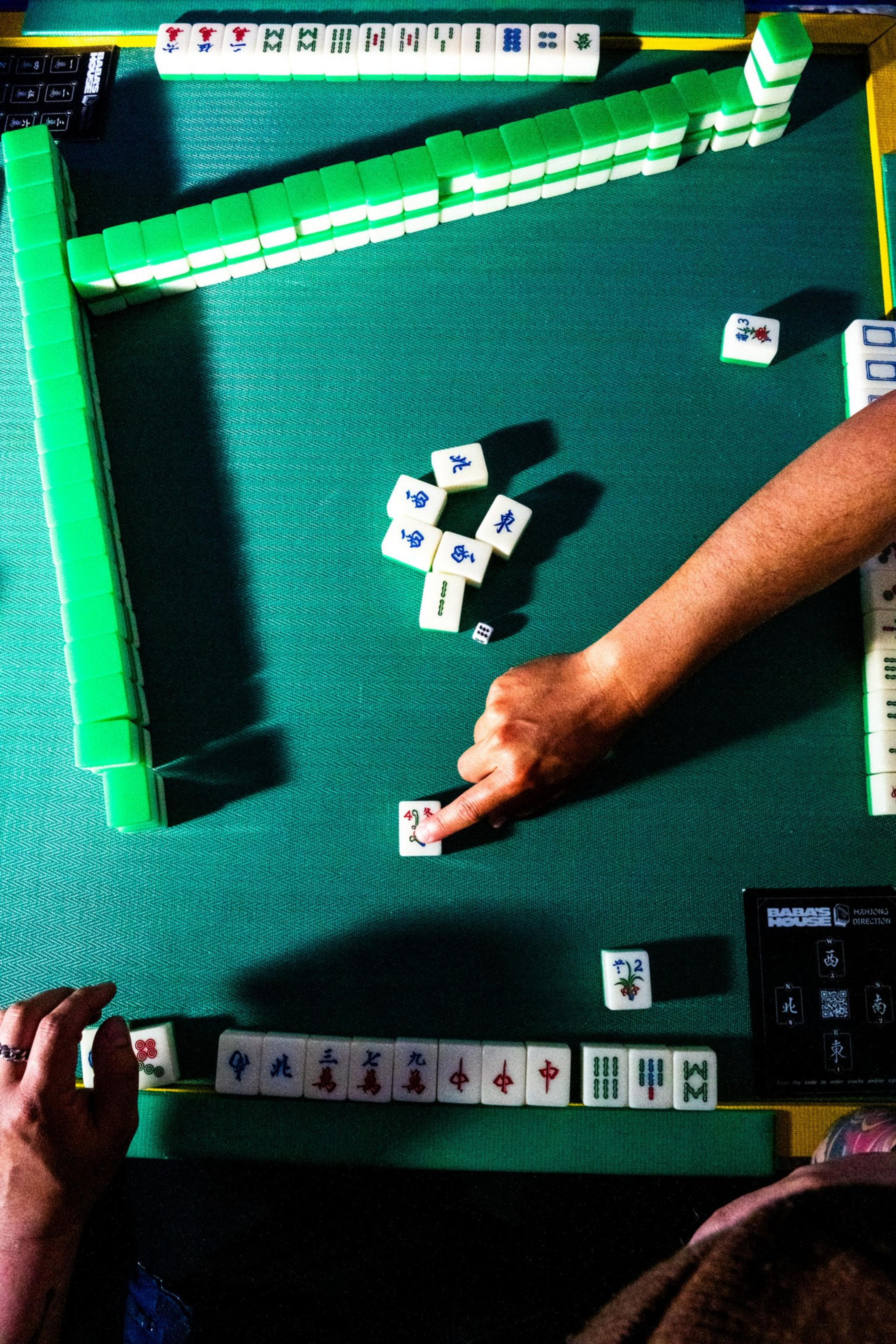 A Mahjong game in progress on a green table, with two players’ hands visible, one player pointing to a tile, and several tiles arranged and stacked in the center.