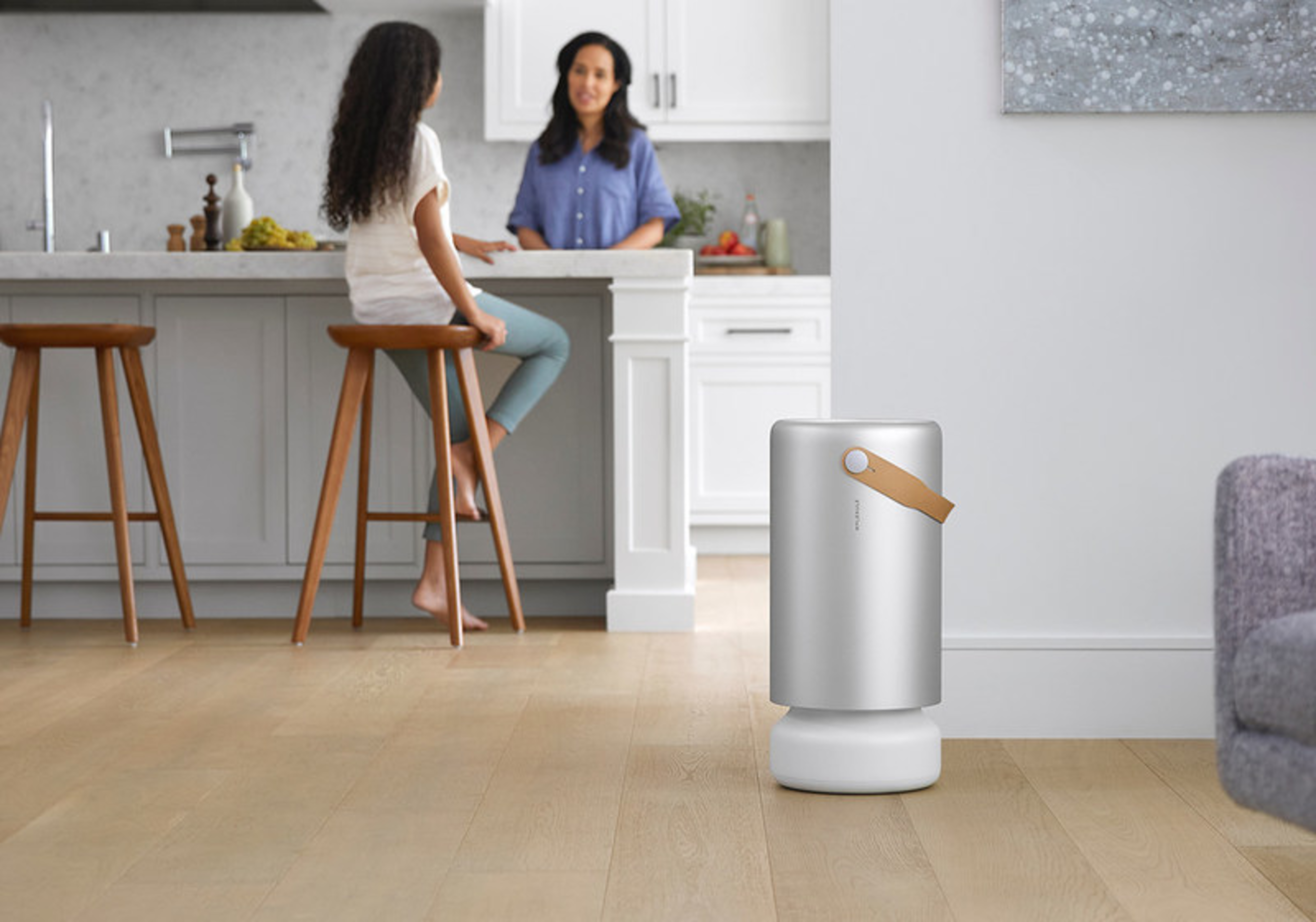 A modern kitchen with two women talking near a counter, and a sleek silver air purifier with a leather strap standing on the wooden floor.