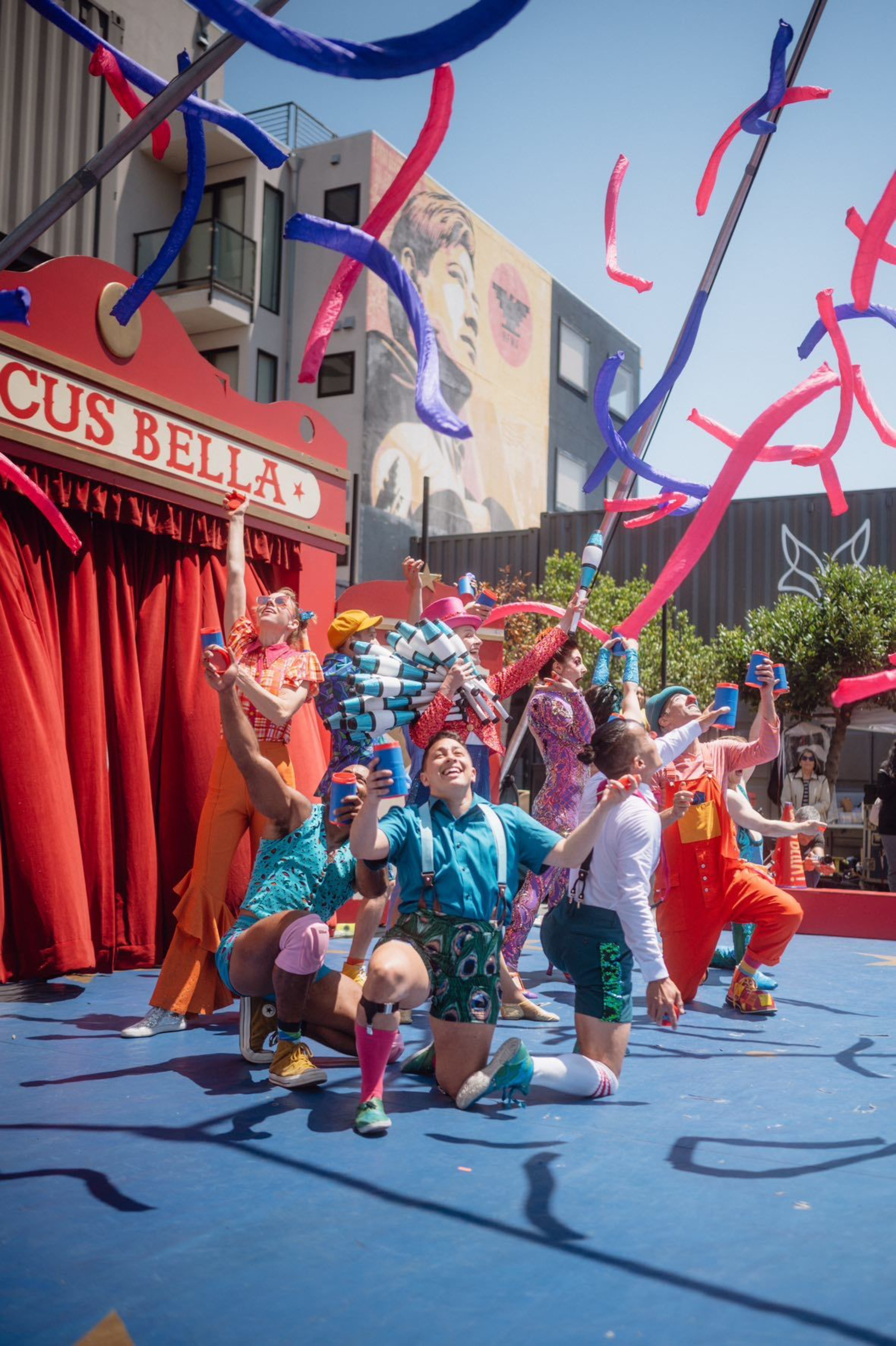 A group of performers in colorful costumes joyfully pose in front of a red circus tent with streamers flying above them, under a clear blue sky.