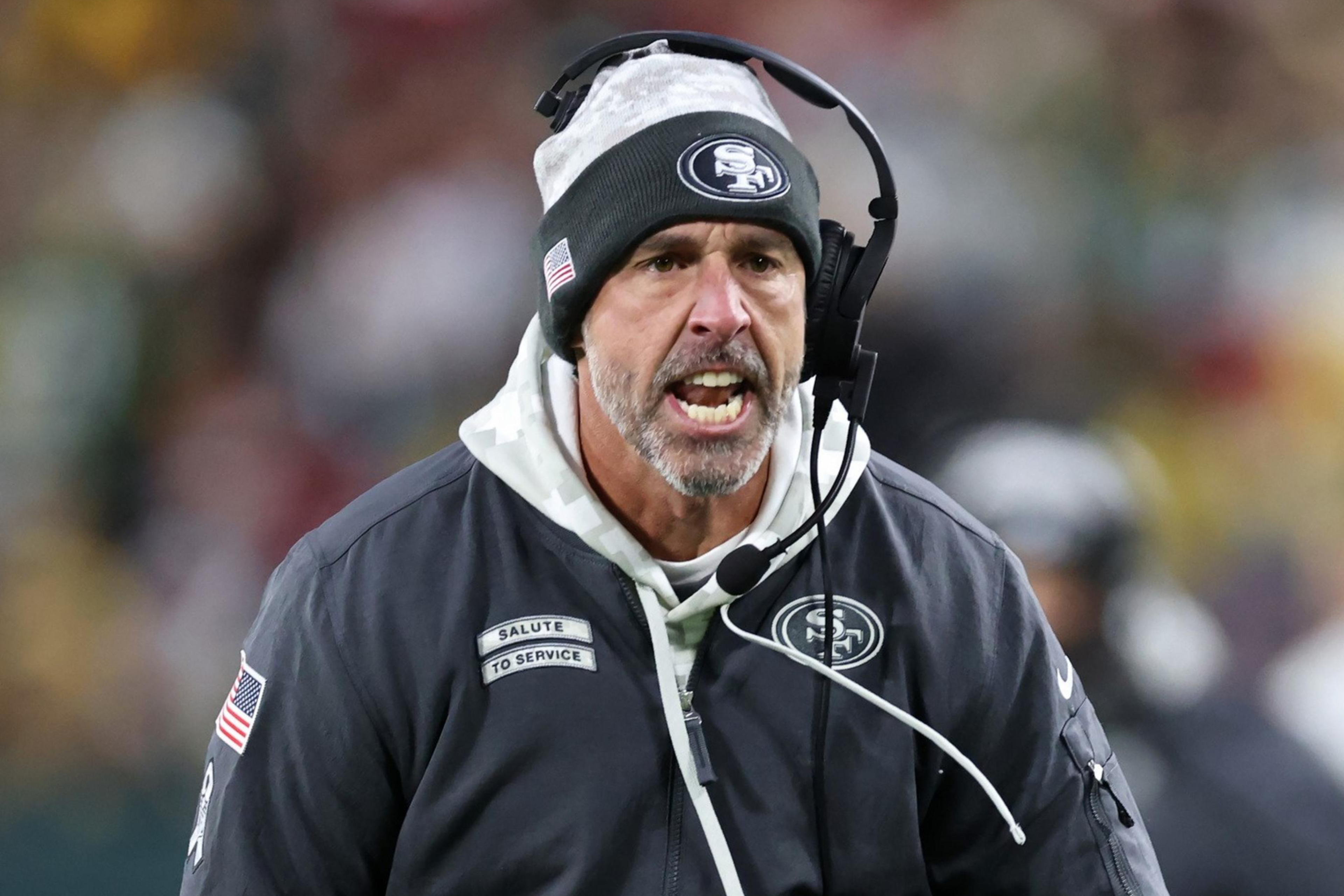 A coach wearing a beanie and headset is on a sports field, holding a folded playbook. He looks intense, with an open mouth, and wears a dark jacket.