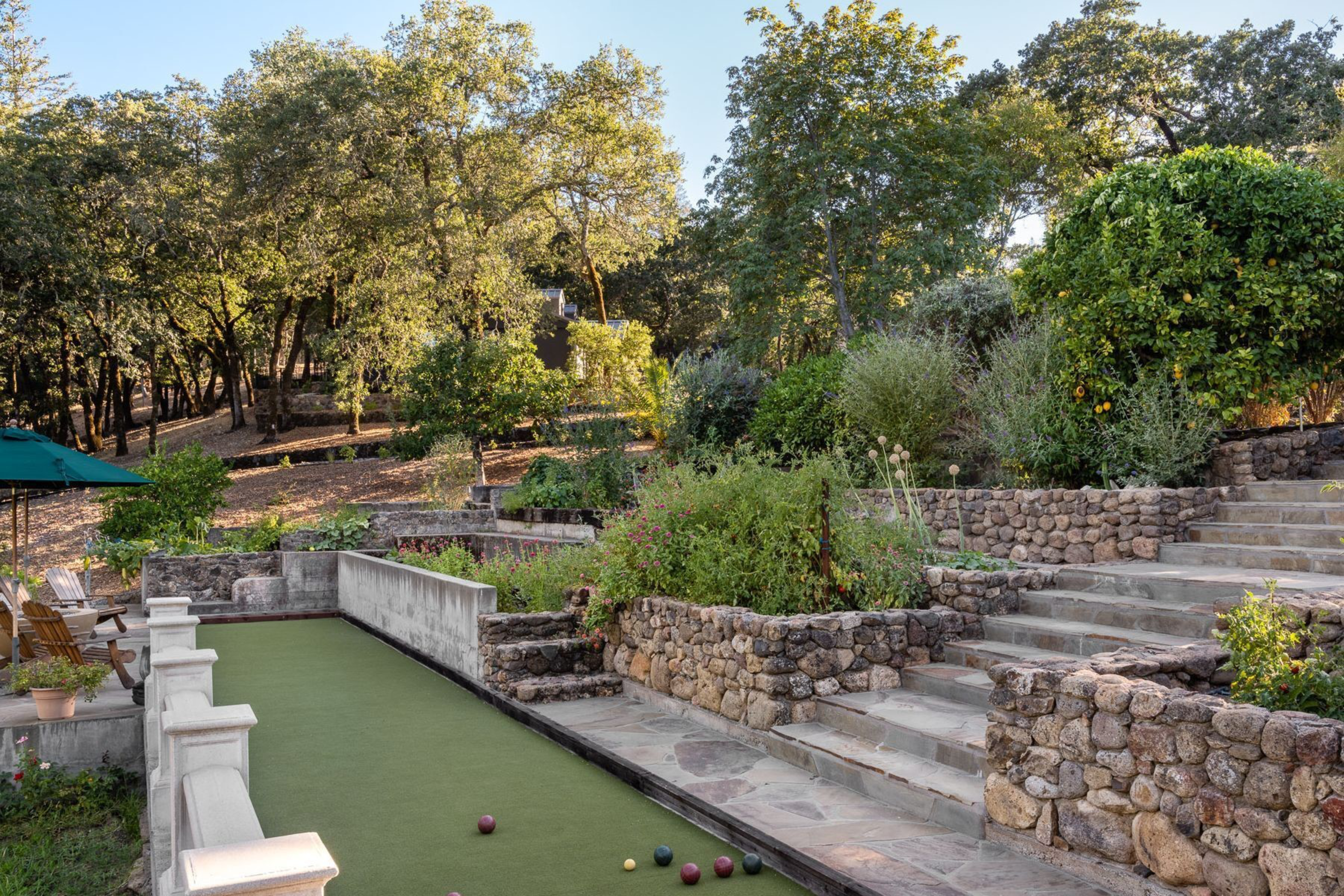 A bocce ball court surrounded by stone steps and lush greenery, with trees and bushes in the background under clear daylight.