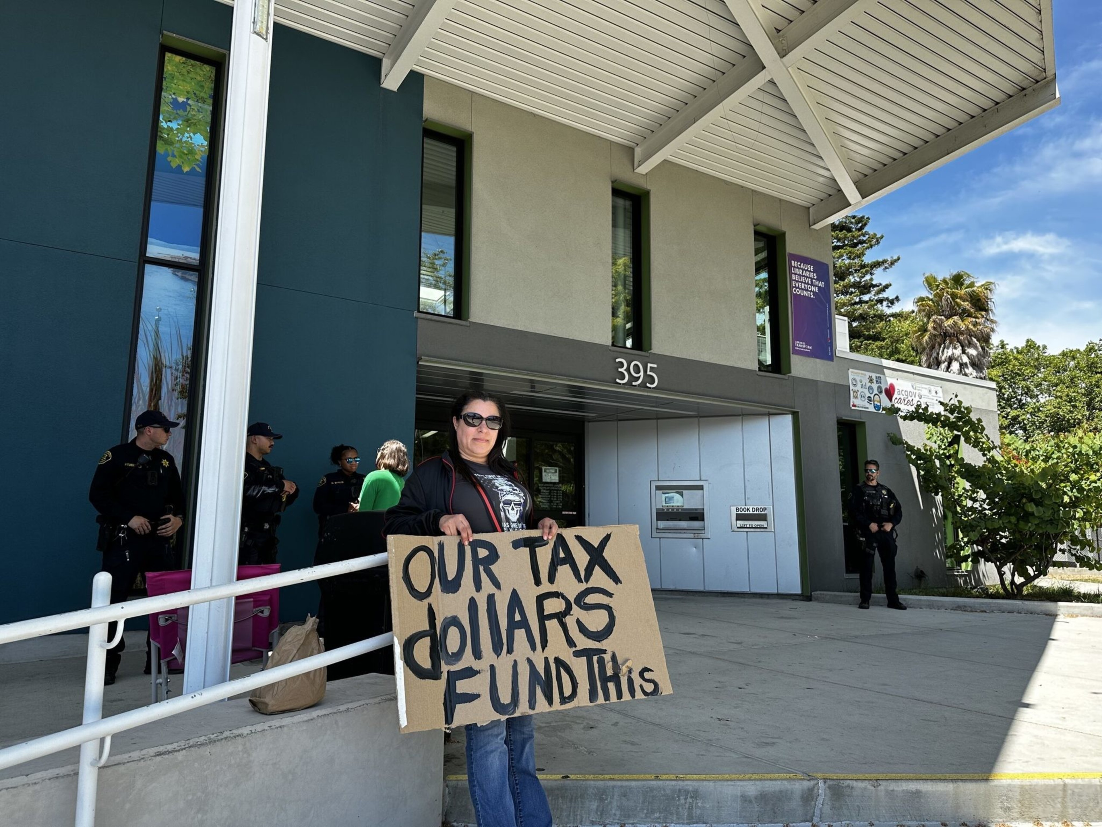 A woman stands outside a building holding a cardboard sign that reads "OUR TAX dollars FUND THIS" with police officers in the background.