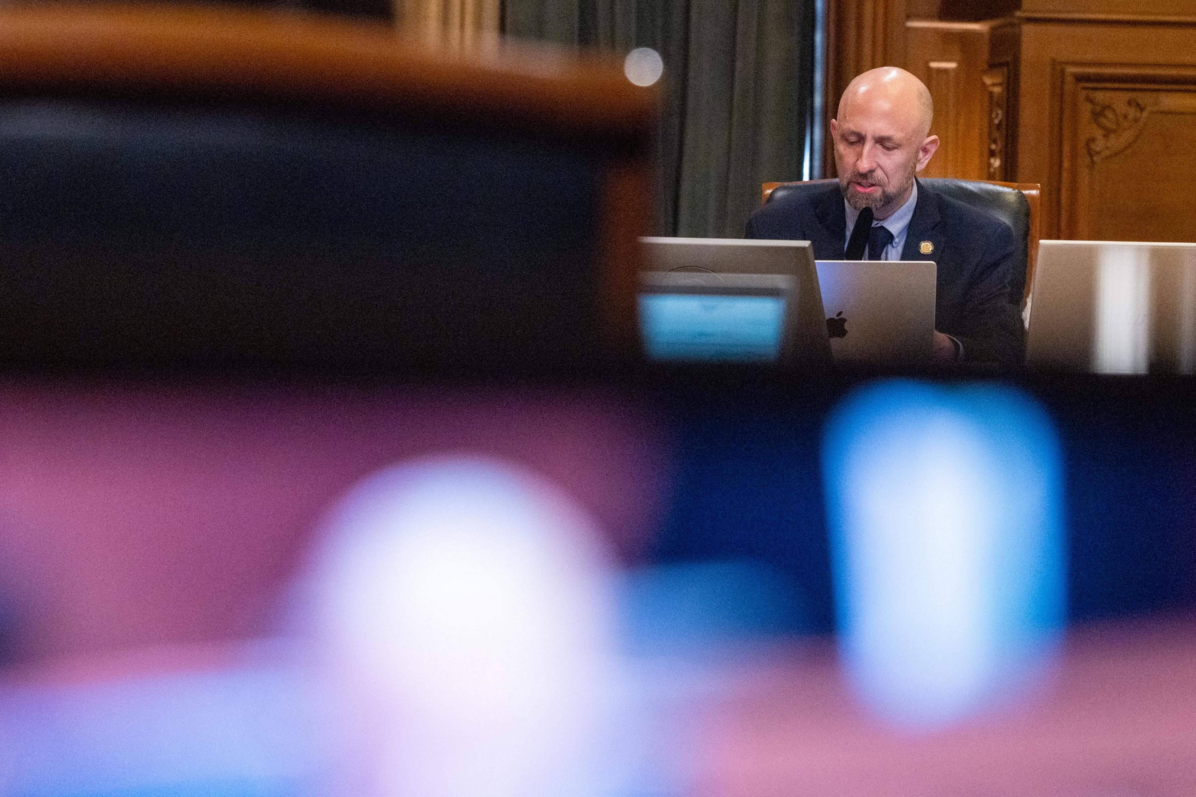 A bald man in a suit sits behind a desk with a laptop, focused on his work, with blurred foreground objects and a wooden wall behind him.