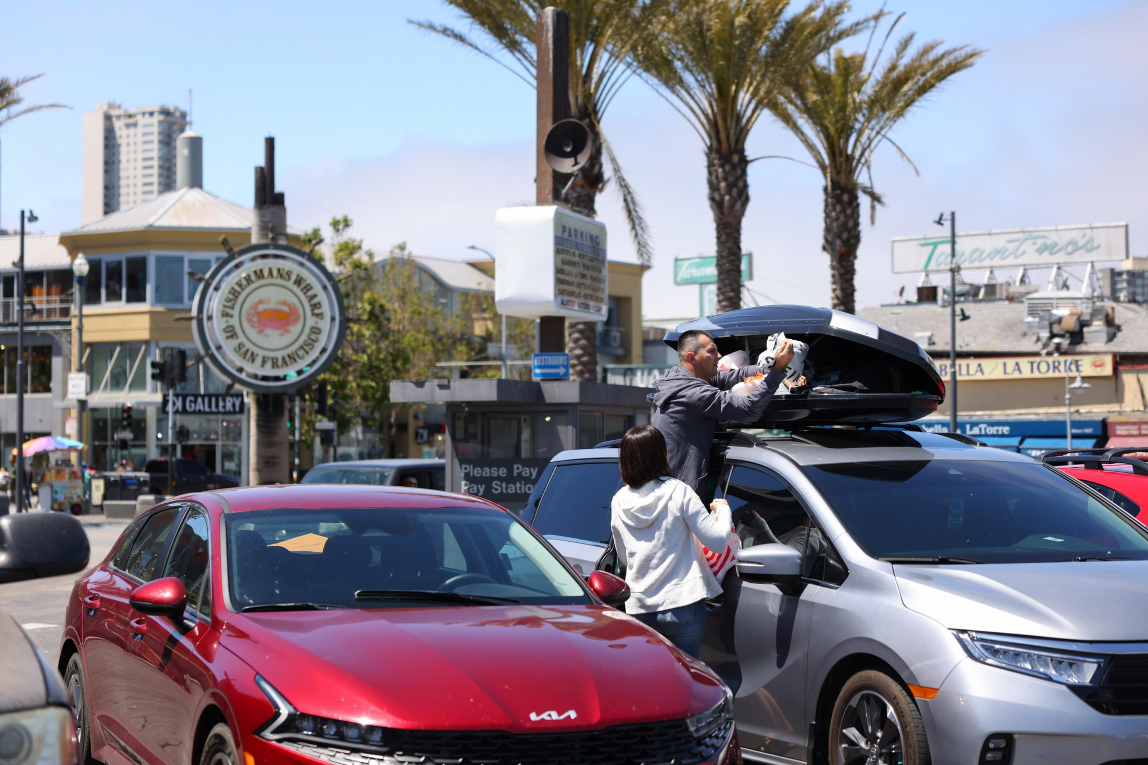 A man and woman load luggage into a rooftop cargo box on a silver SUV parked near palm trees and a Fisherman’s Wharf sign in San Francisco.