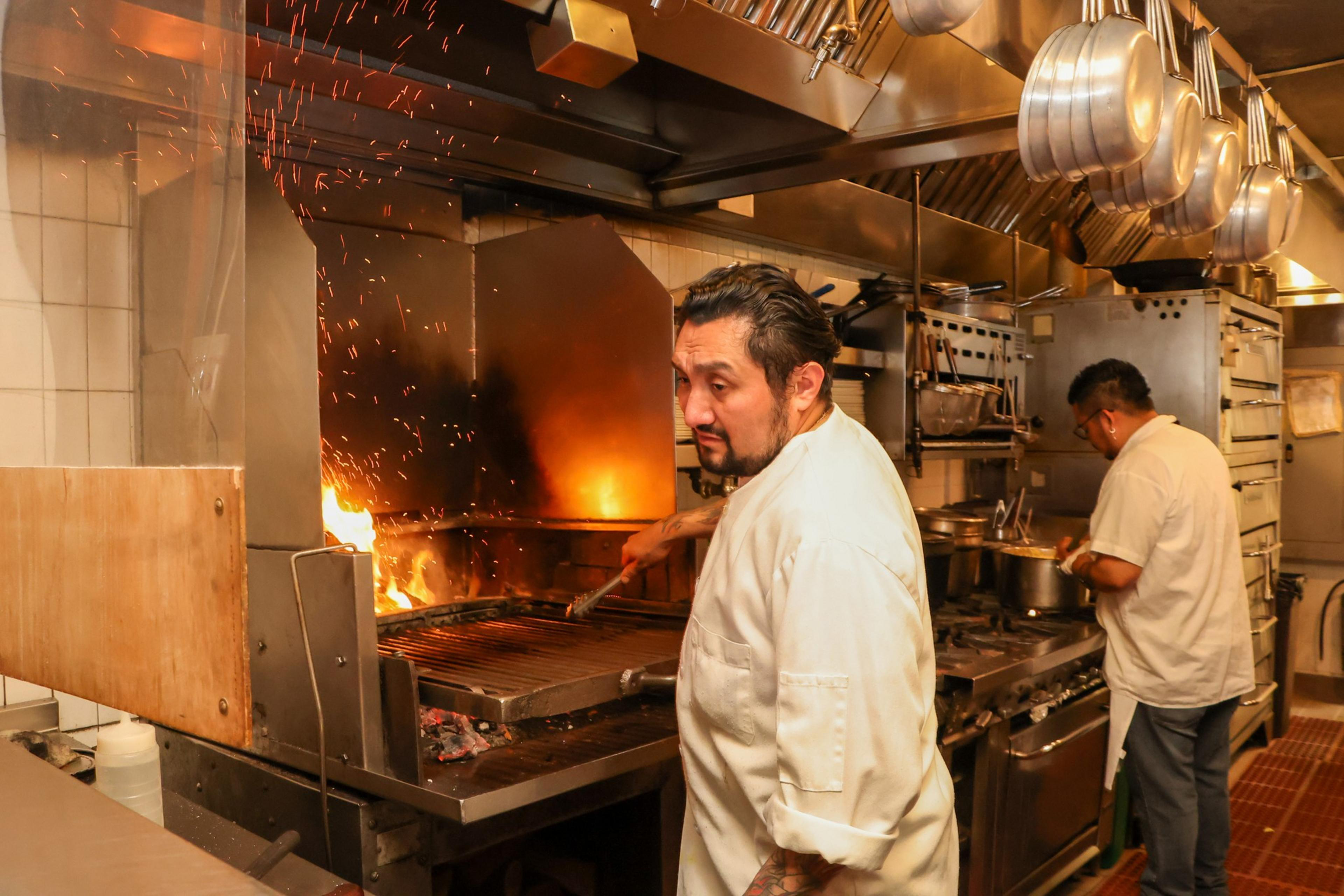 A chef in a white coat uses tongs to grill over an open flame with sparks flying, while another person works at a stove in a commercial kitchen.