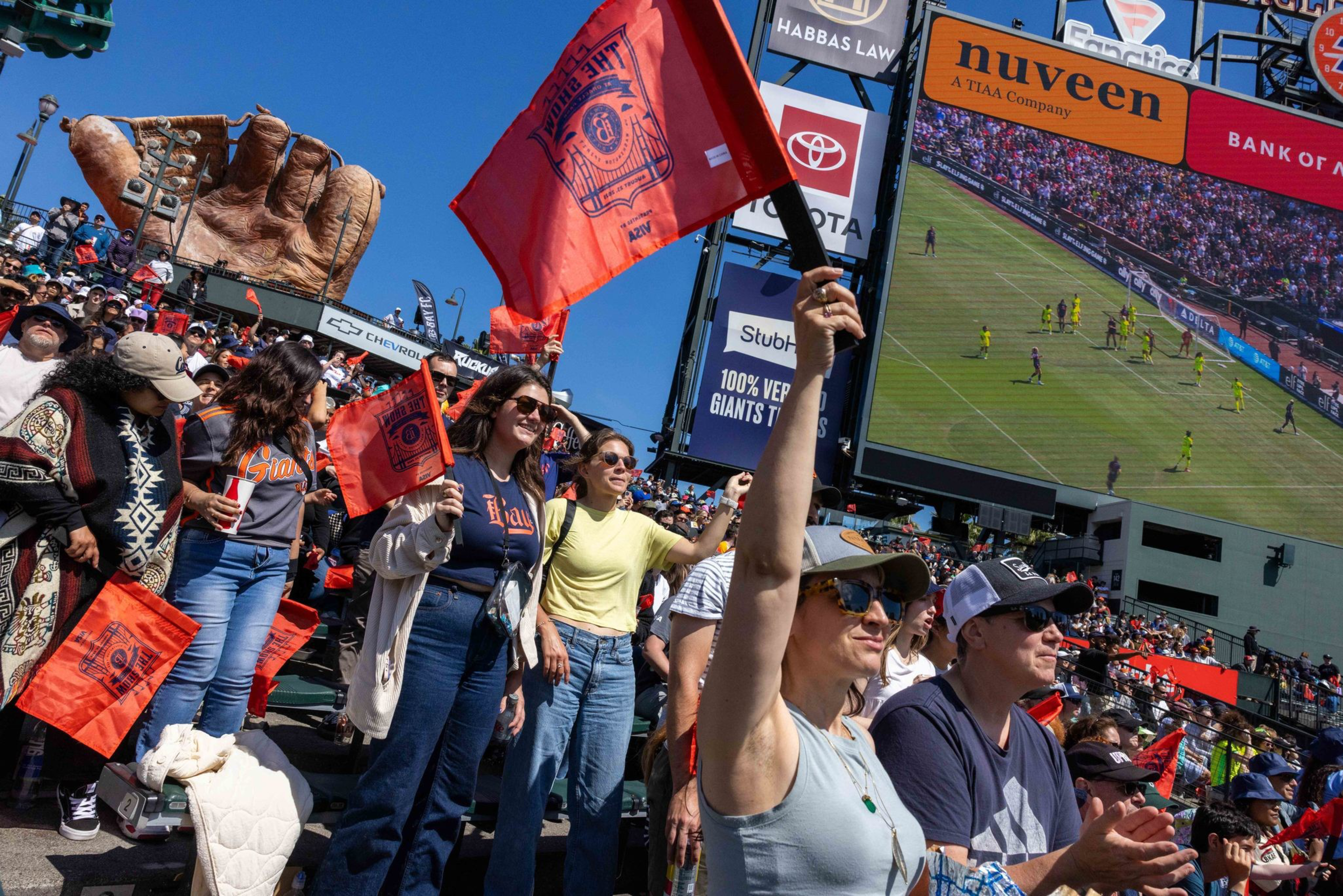 Fans cheer while waving orange flags at an outdoor stadium during a soccer match shown on a large screen under a sunny blue sky.