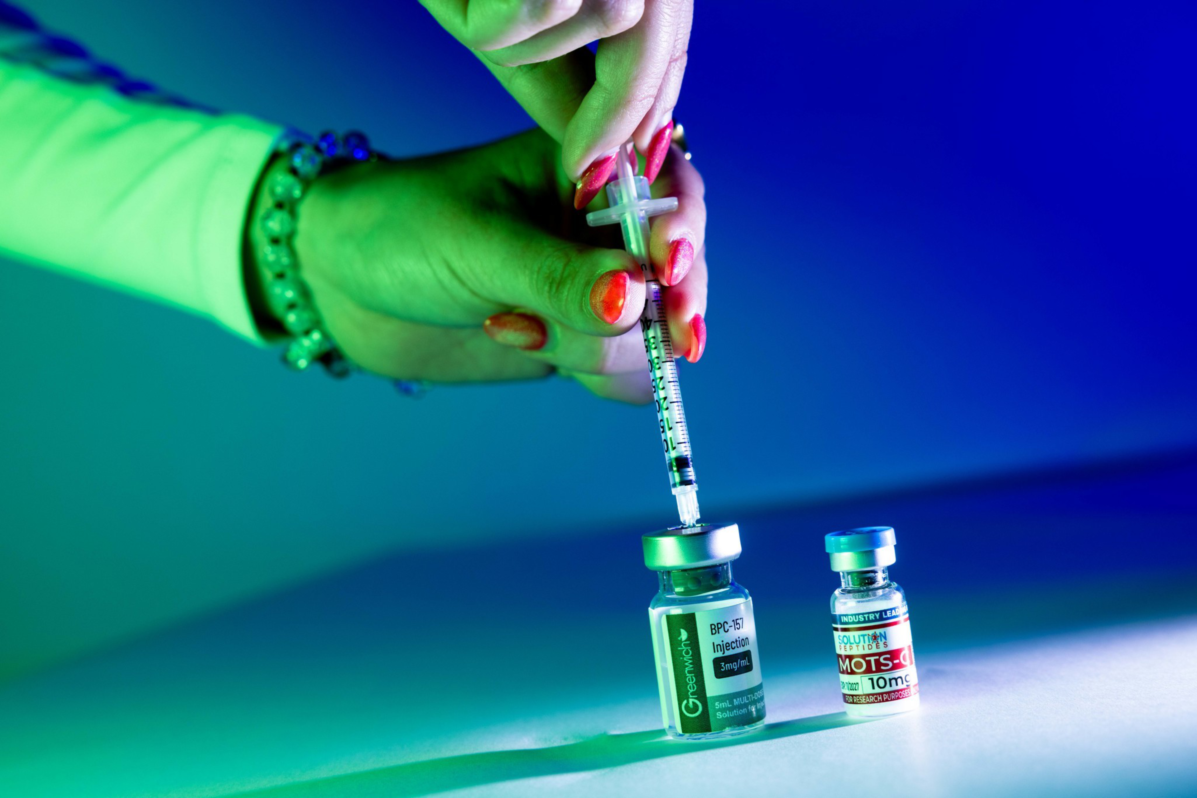 A hand with red-painted nails holds a syringe drawing liquid from a vial labeled “BPC-157 Injection” beside another vial labeled “MOTS-c.”