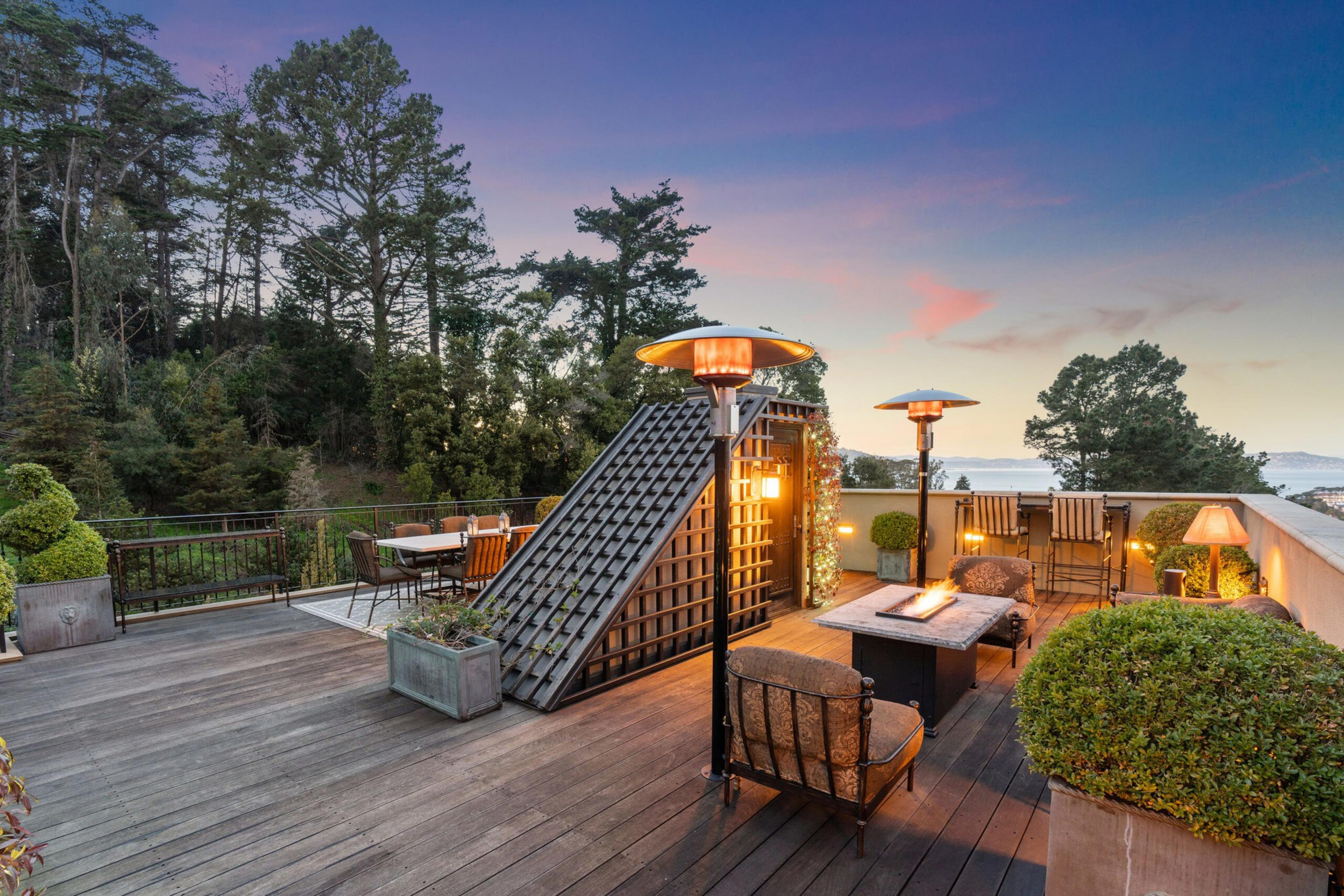 A spacious rooftop patio at dusk features wooden decking, outdoor seating with cushions, two heaters, potted plants, and a view of trees and distant water.