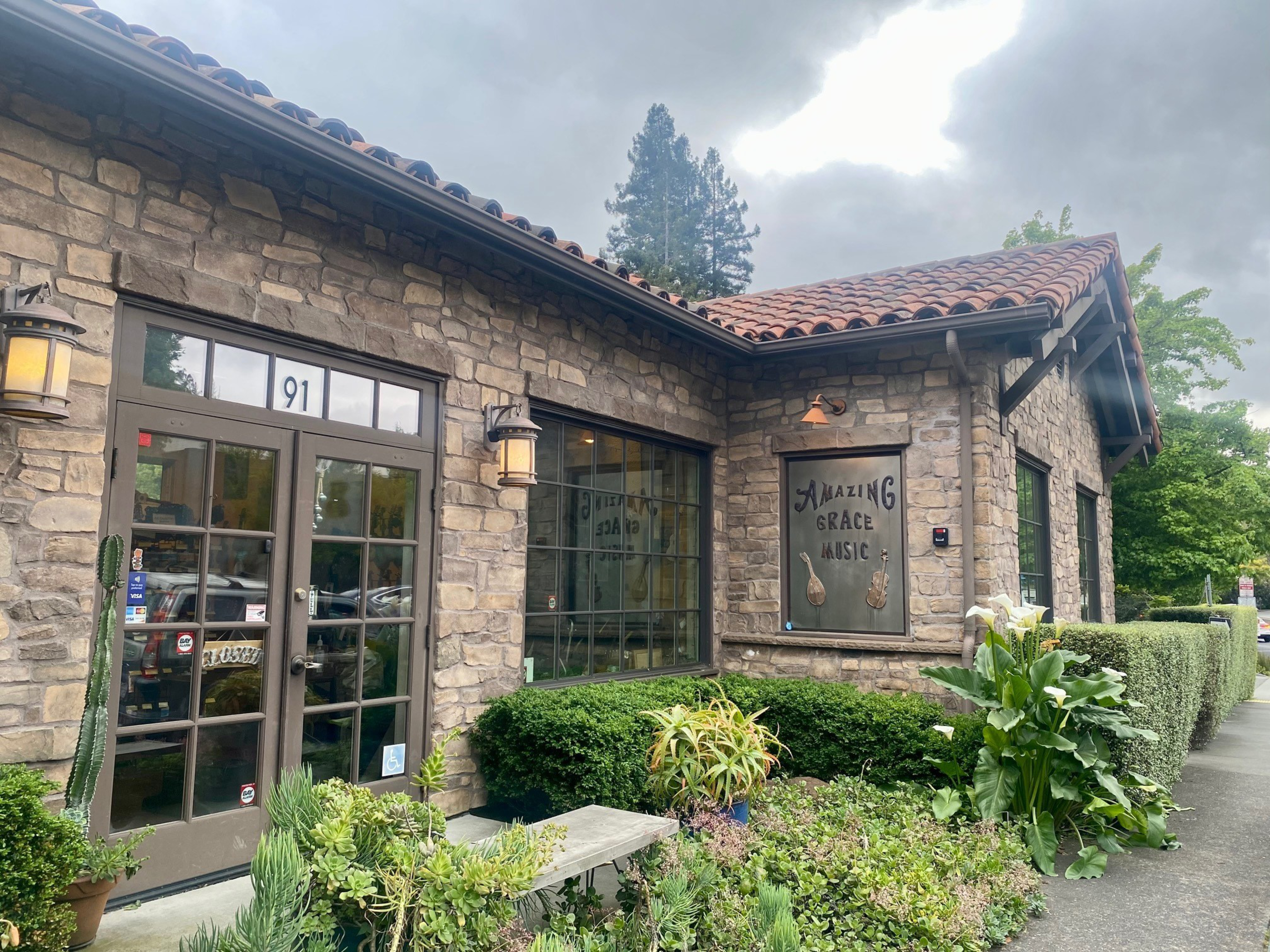 A stone building with a red tiled roof houses Amazing Grace Music, surrounded by lush green plants and shrubs under a cloudy sky.