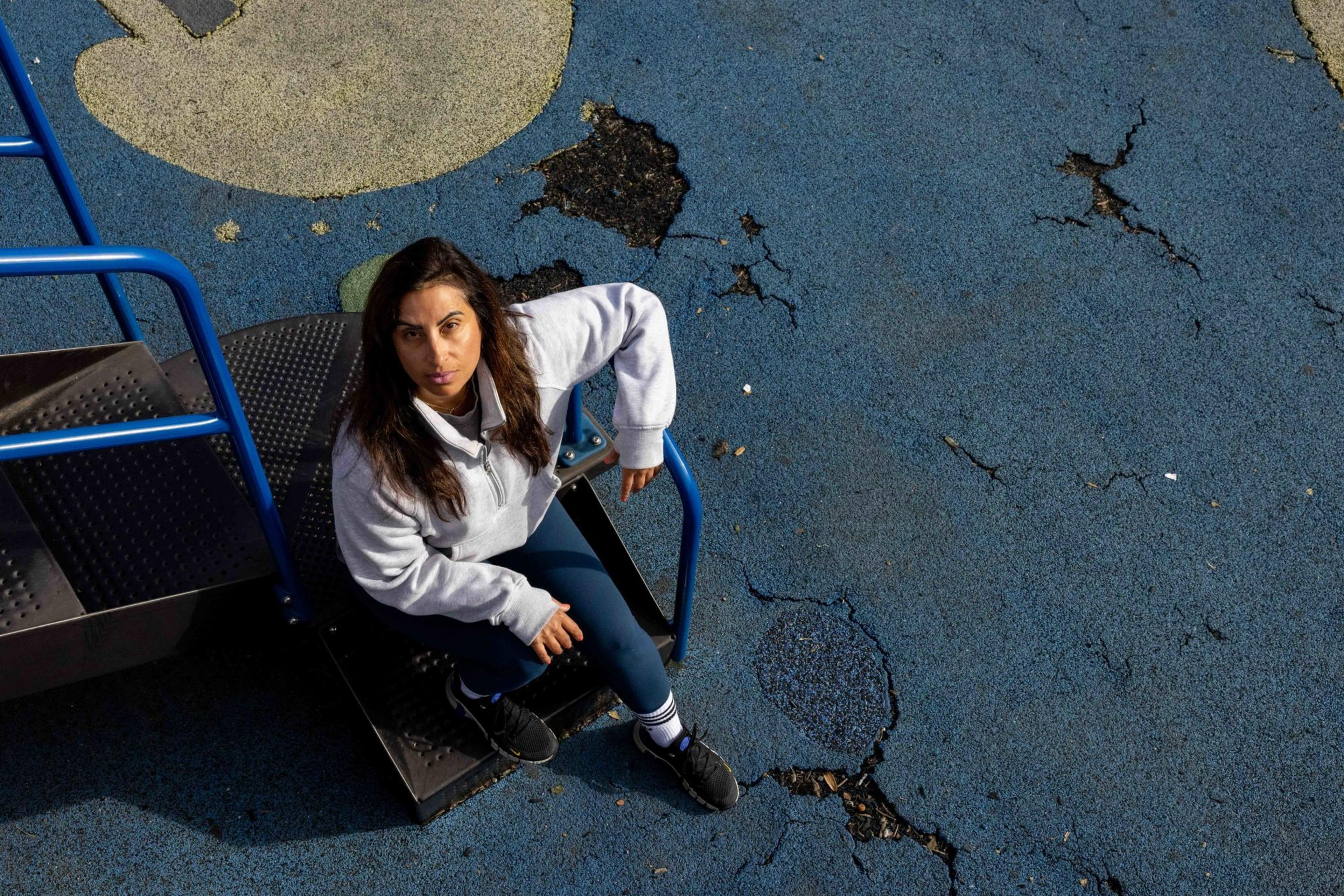 A woman sits on playground steps, looking up towards the camera. She's wearing a light sweatshirt and dark leggings. The ground around her is blue and cracked.