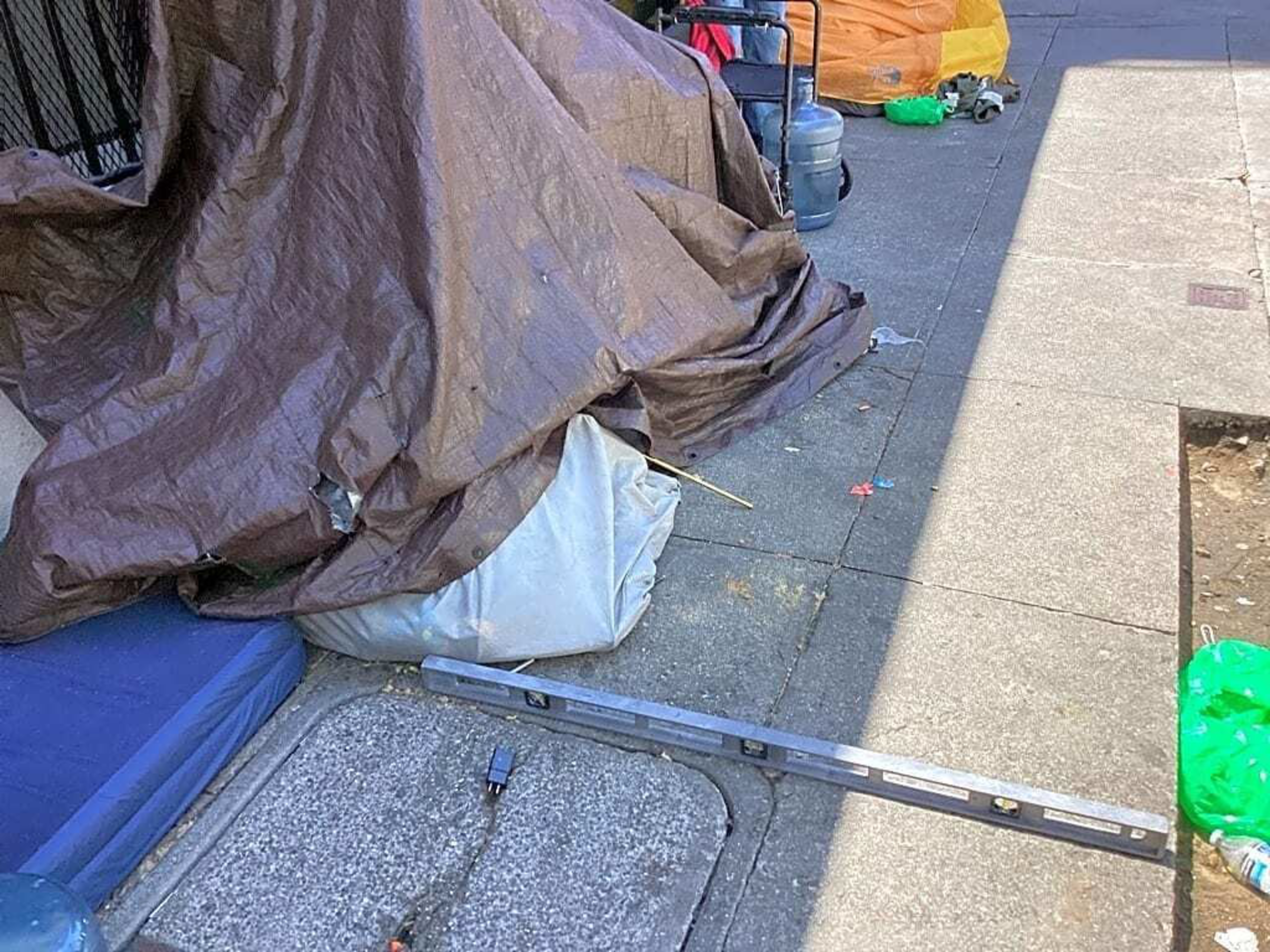 A street scene with a makeshift tent covered by a brown tarp, surrounded by various items like a blue mattress, a long level tool, bottles, and other belongings.