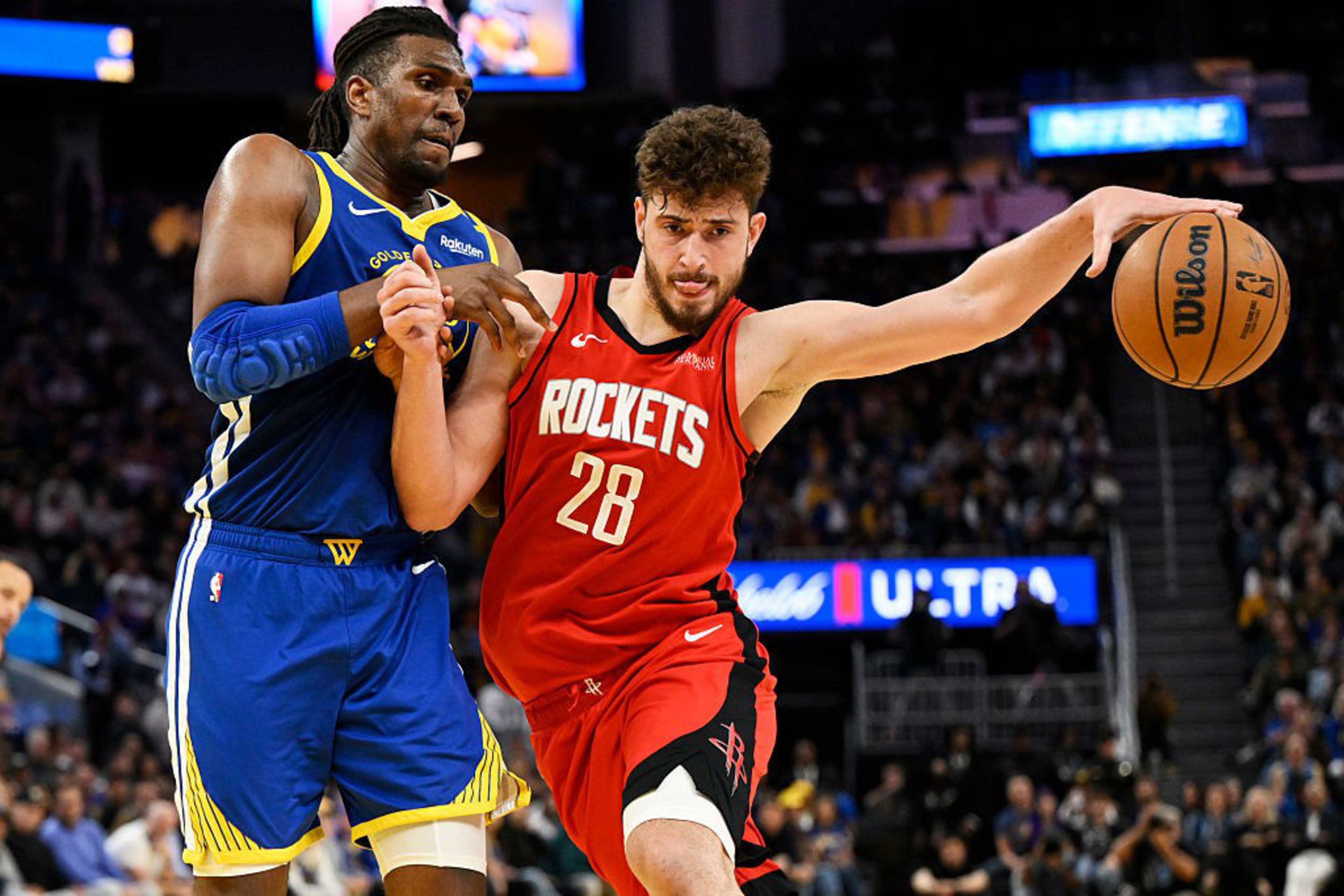 Two basketball players are in action on the court. One player in a blue jersey is defending, while the other in a red jersey is holding the ball.