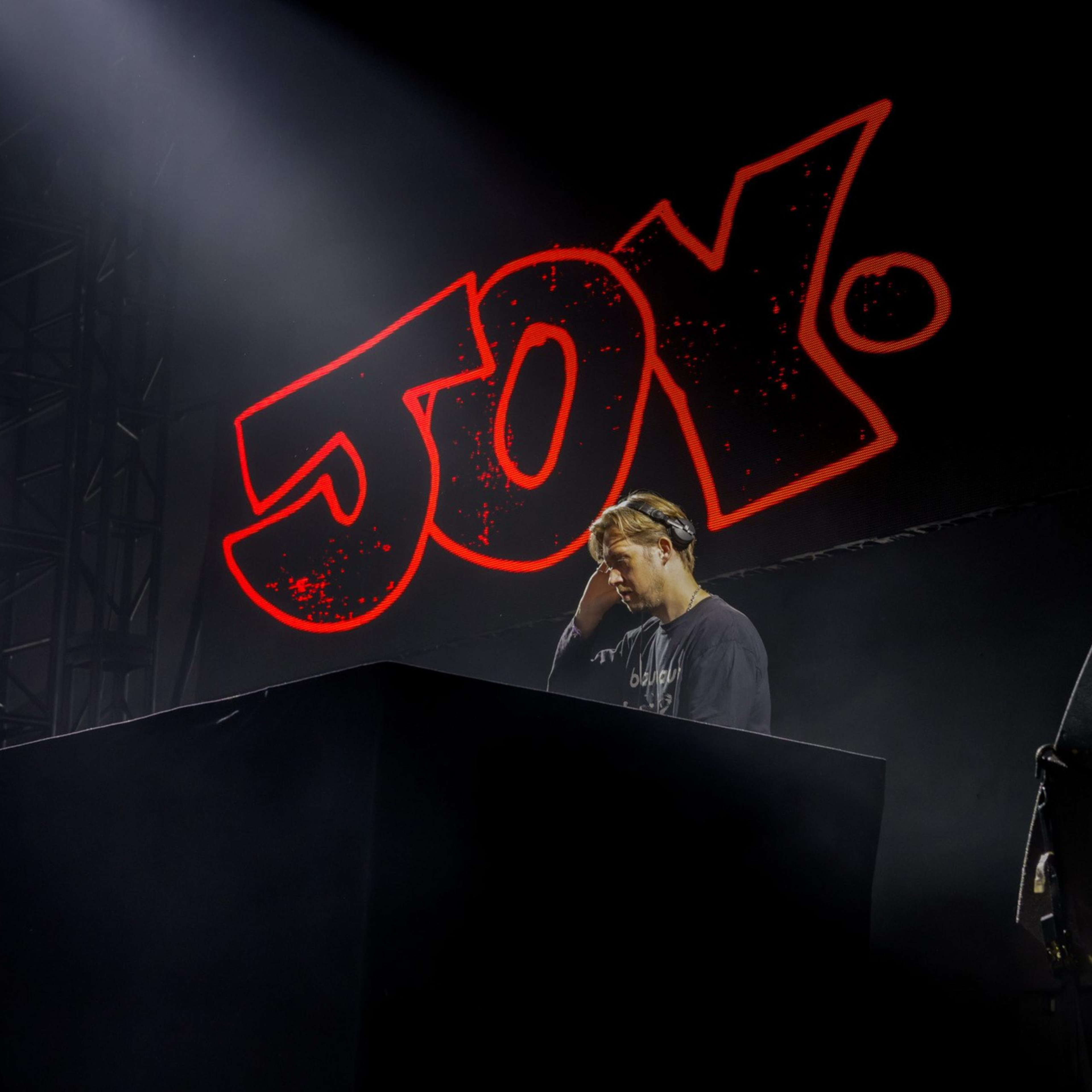 A DJ stands focused at his turntable, illuminated under stage lights, with the word &quot;Joy&quot; in large, bright red letters behind him.
