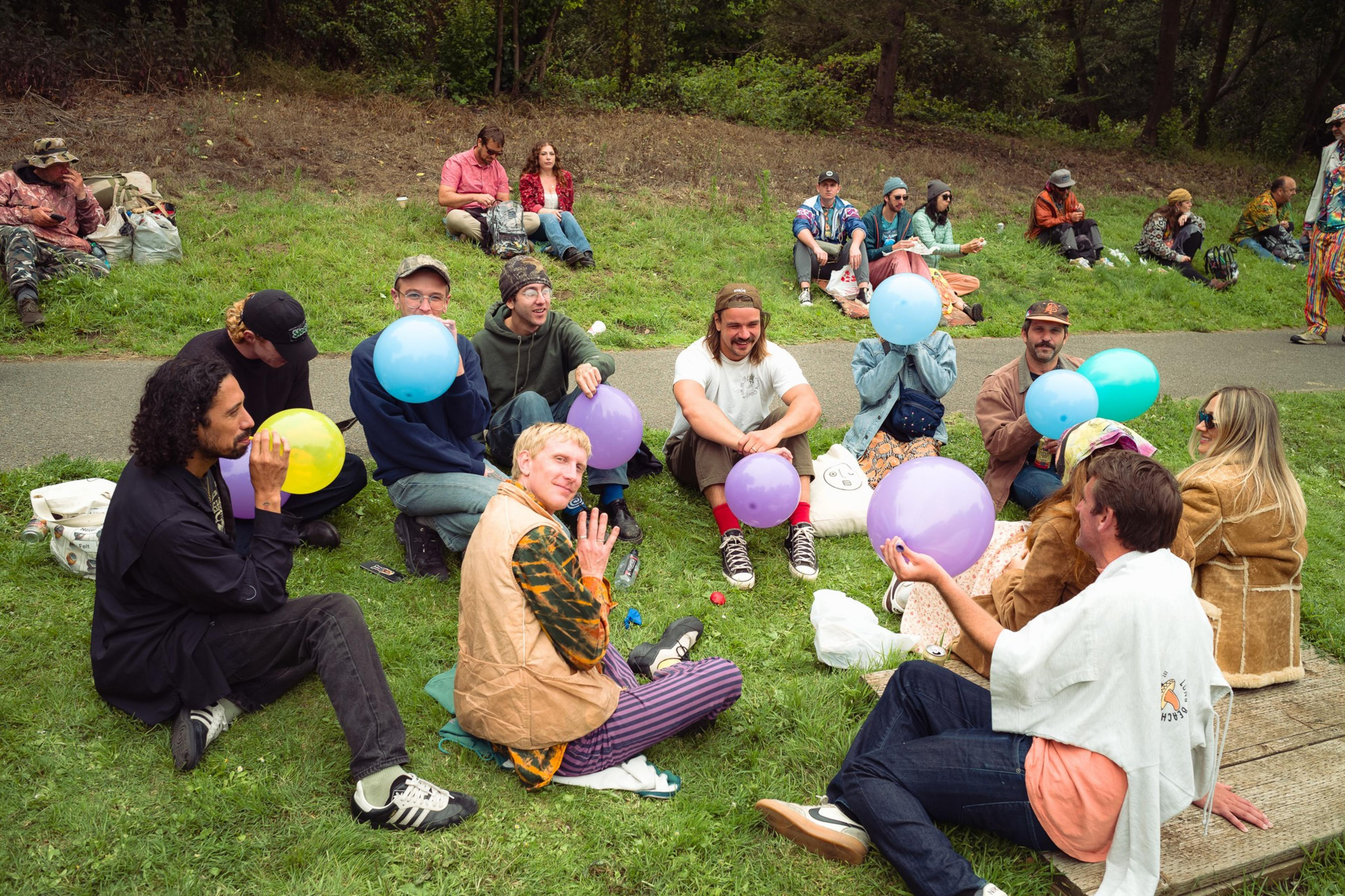 A group of people sit on grass, holding colorful balloons, with others sitting on a hill and a path behind them in a park-like setting.