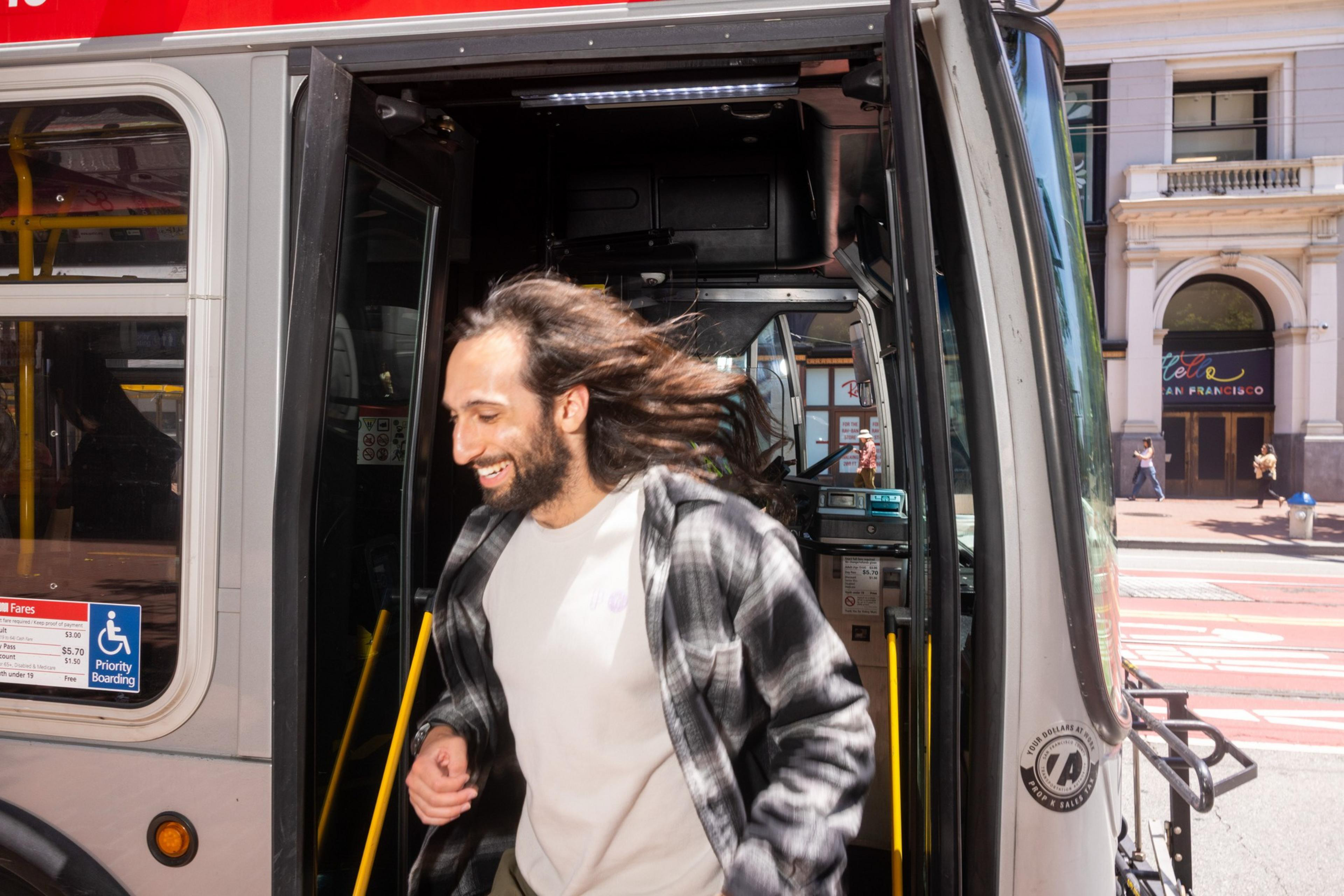 A smiling man with long hair and a plaid shirt is exiting a bus on a sunny street. A building with &quot;Francisco&quot; in the window is visible in the background.