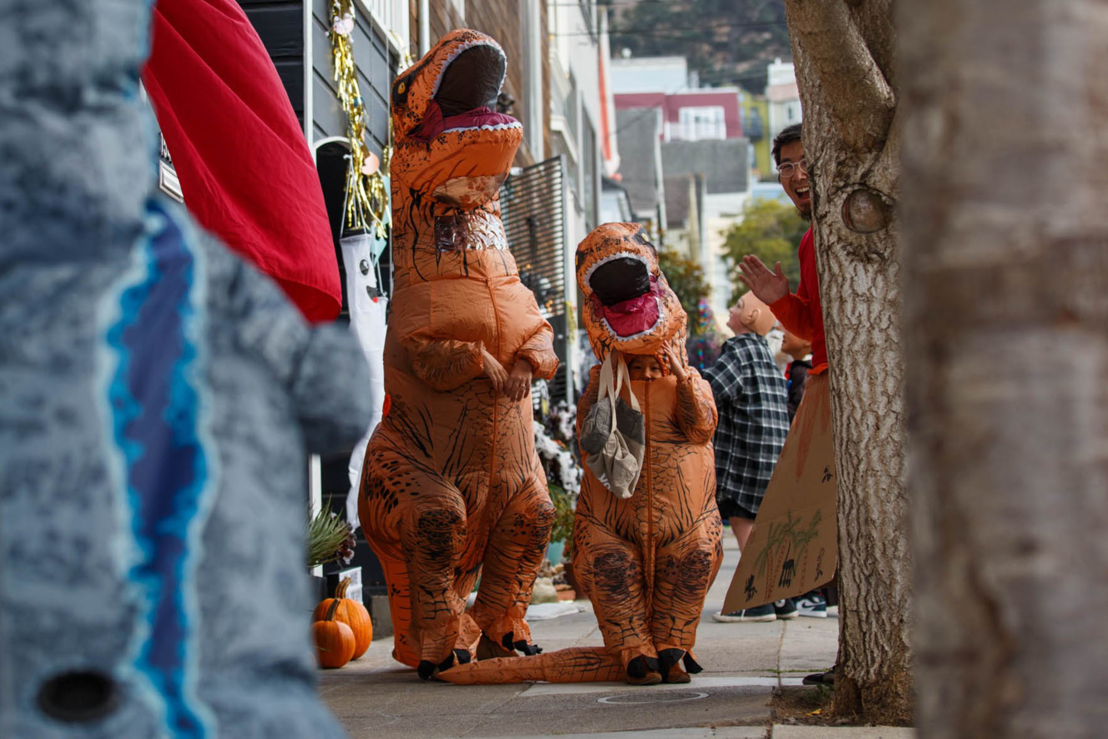 Two people in inflatable orange T-Rex costumes stand on a sidewalk, as a person in a blue dinosaur costume and others watch nearby.