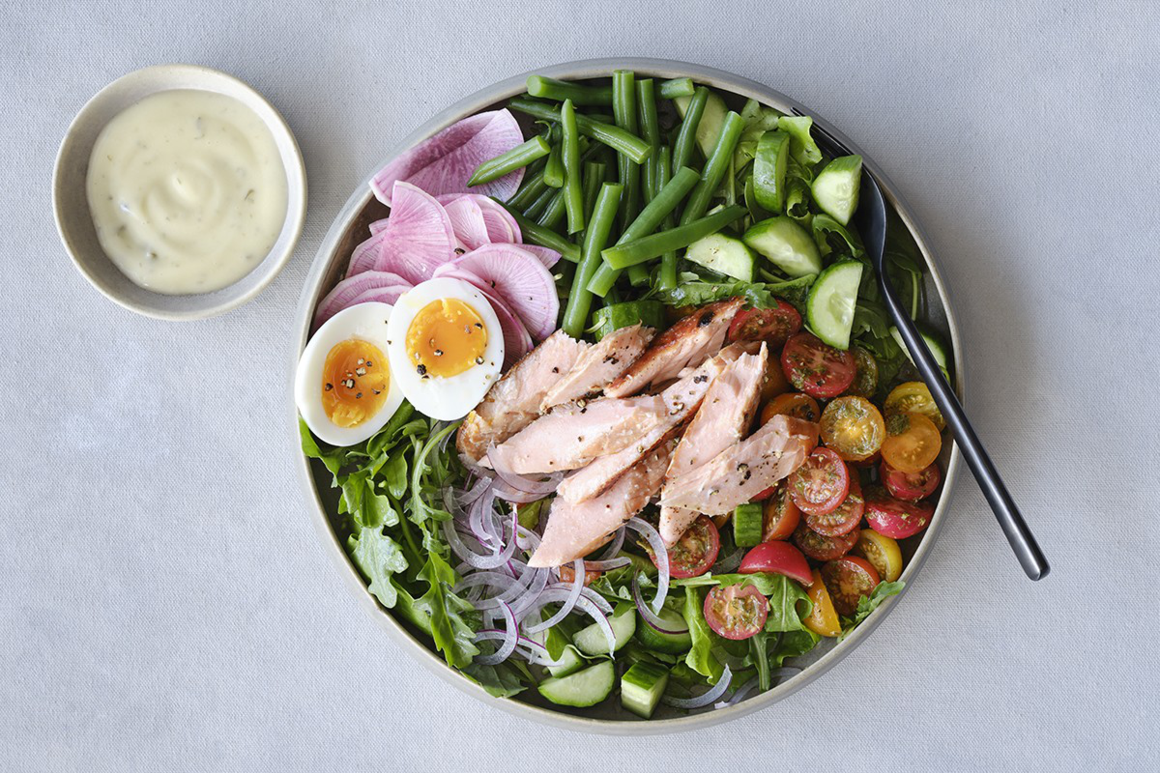 Grilled salmon, green beans, sliced cucumbers, cherry tomatoes, radish, red onion, soft-boiled egg, and mixed greens are arranged on a plate with a side of creamy dressing.