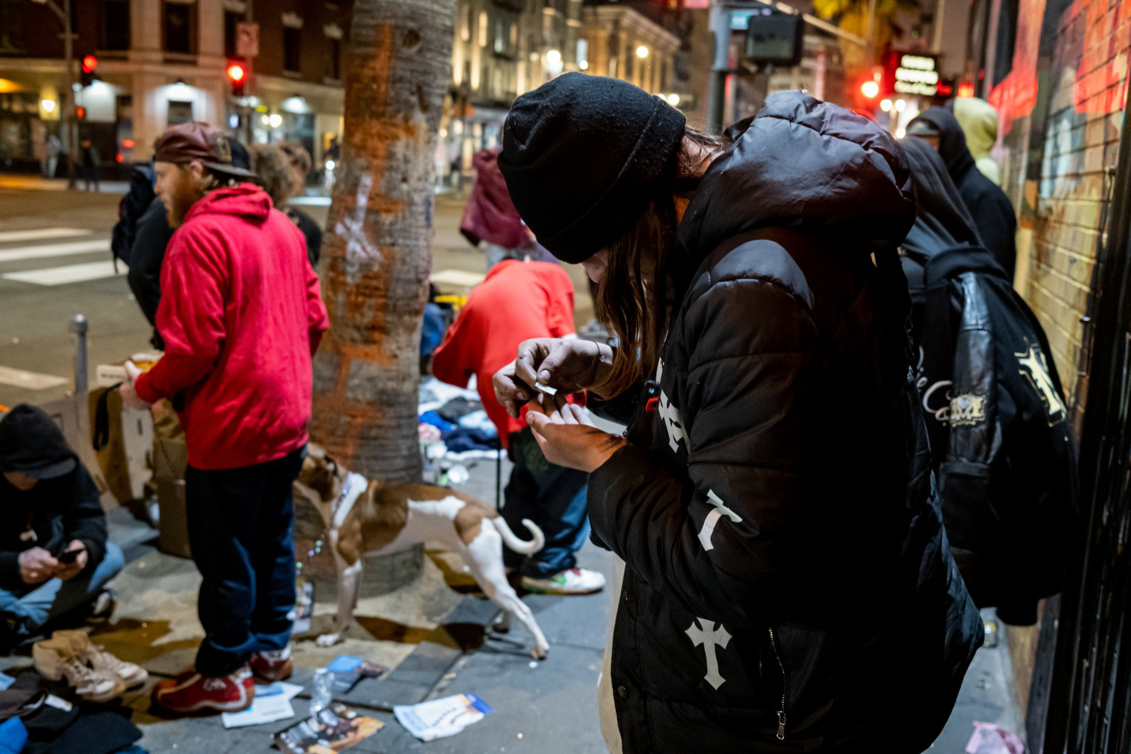 A street scene at night features several people, some standing, others sitting on the ground. A dog stands nearby. One person focuses on an object in their hands.