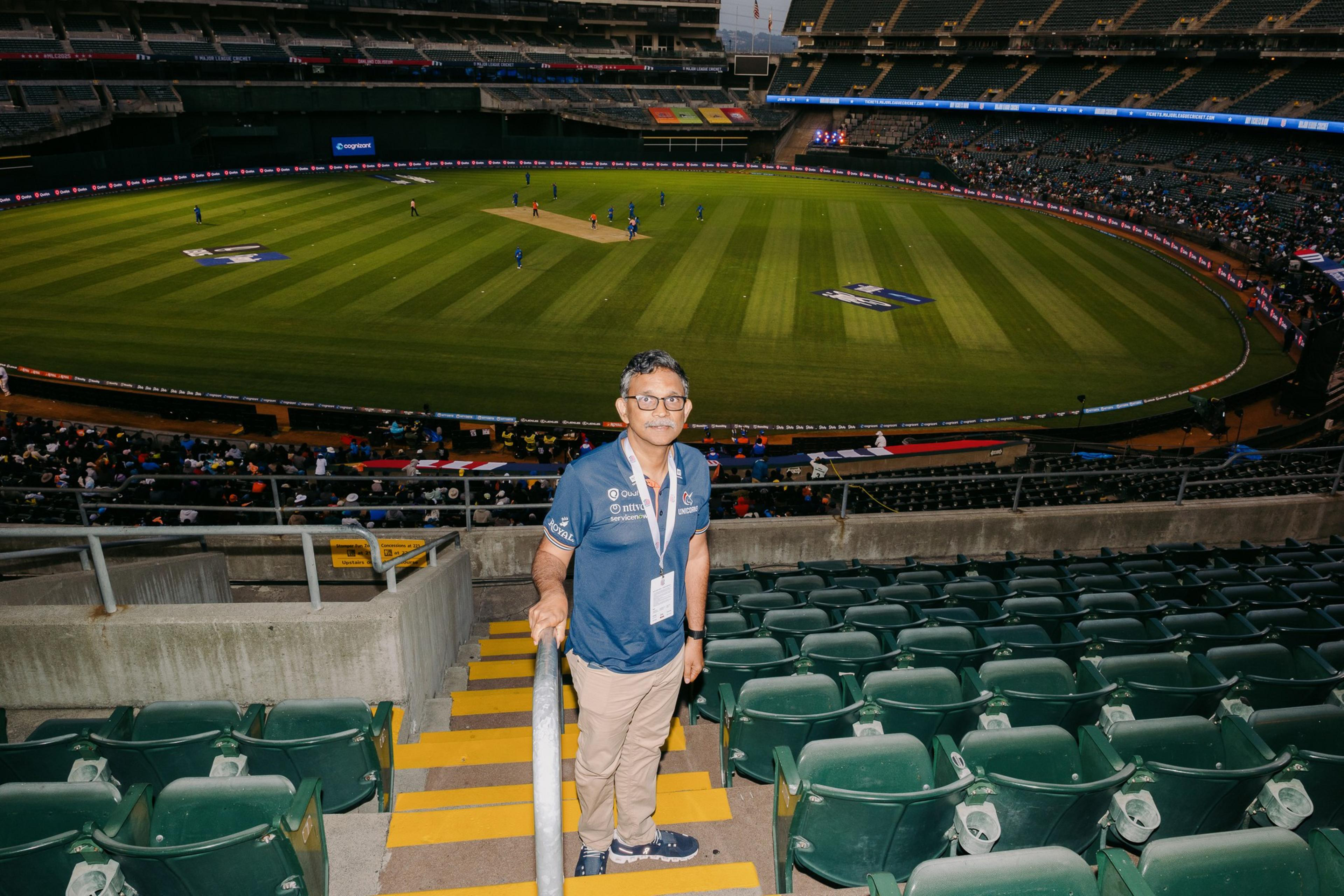 A person stands on stadium stairs, with a cricket field in the background. Some players are on the field, and spectators are in the stands.