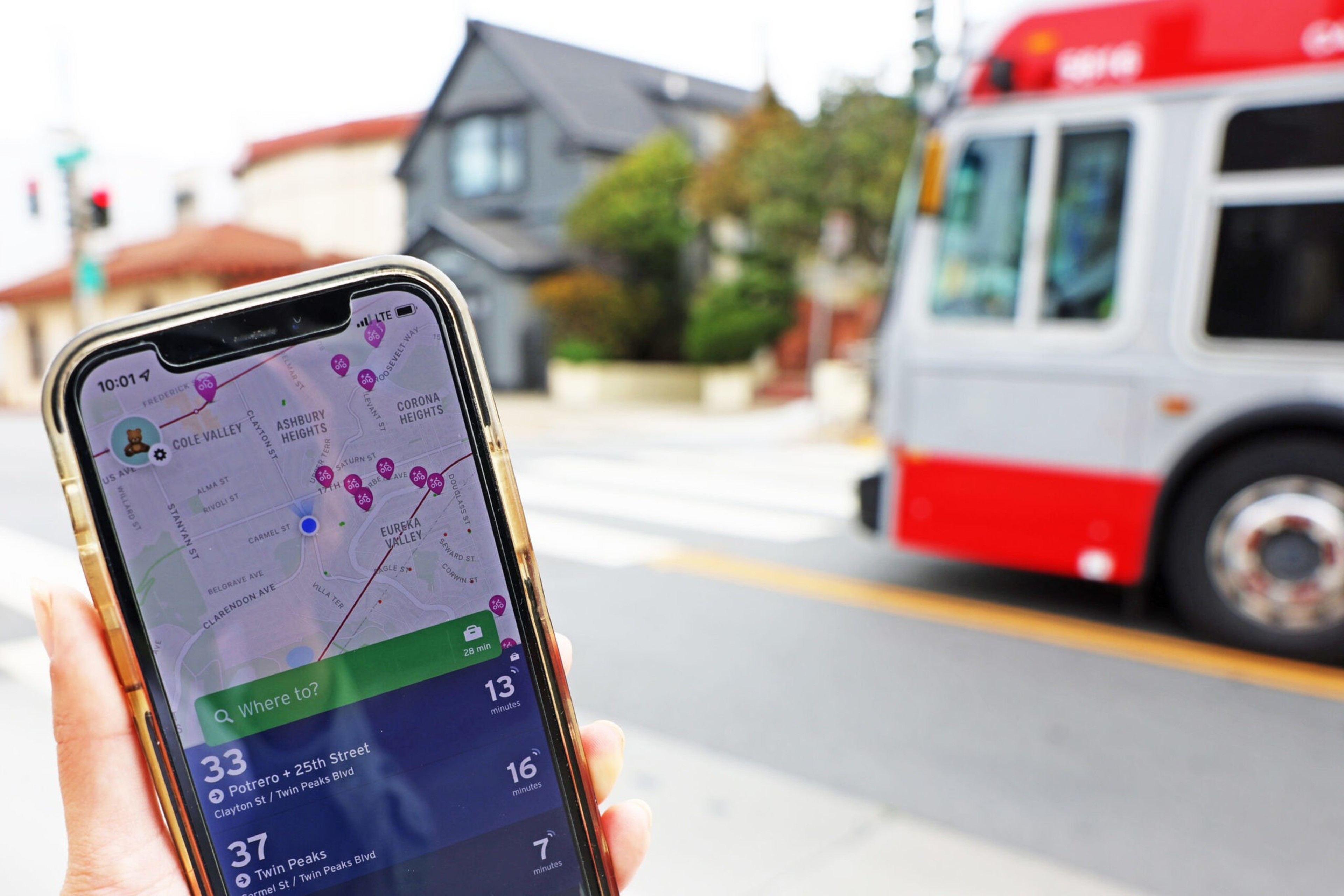 A hand holds a phone showing a transit app with bus routes; a red and white bus passes on the street near houses in the background.