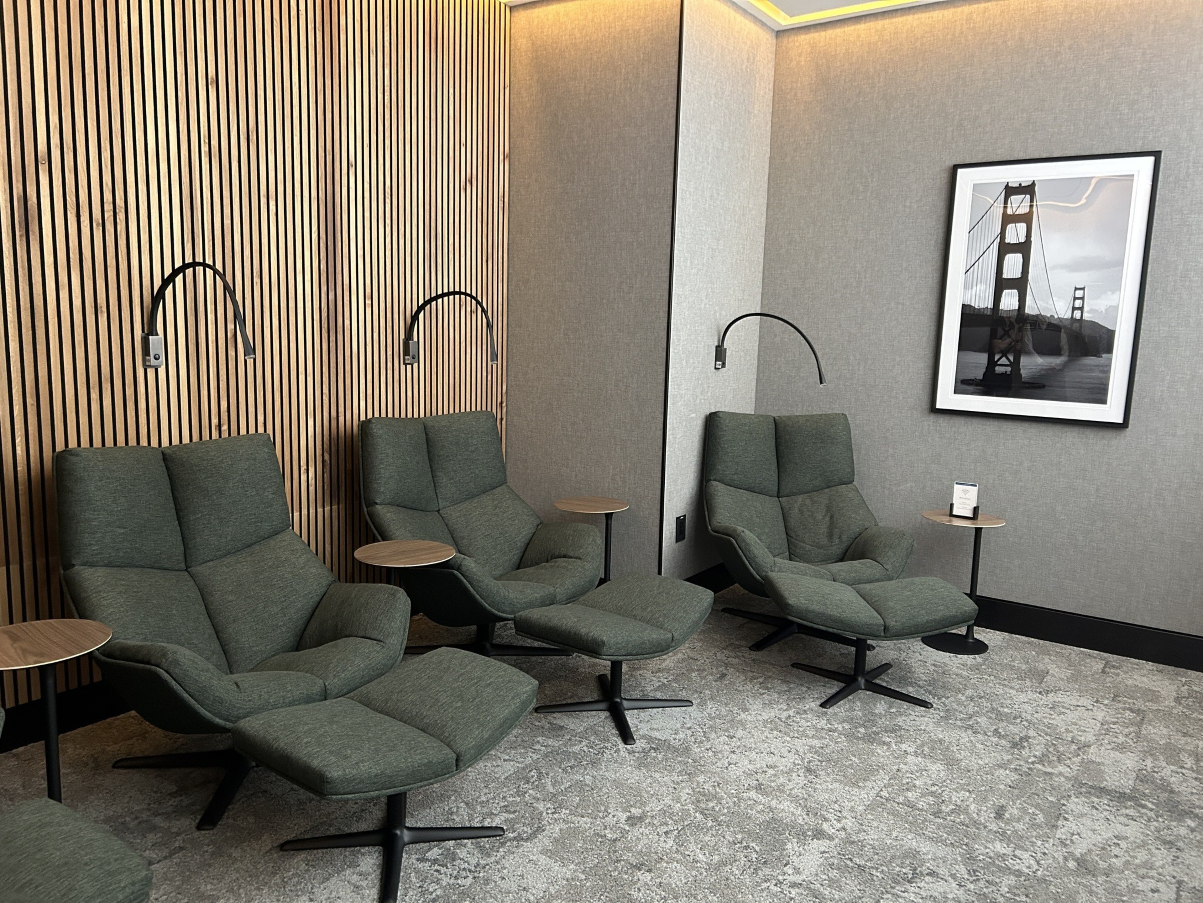 The image shows three green recliner chairs with footrests in a modern lounge, featuring wooden slat paneling, side tables, overhead reading lights, and a framed picture of a bridge.
