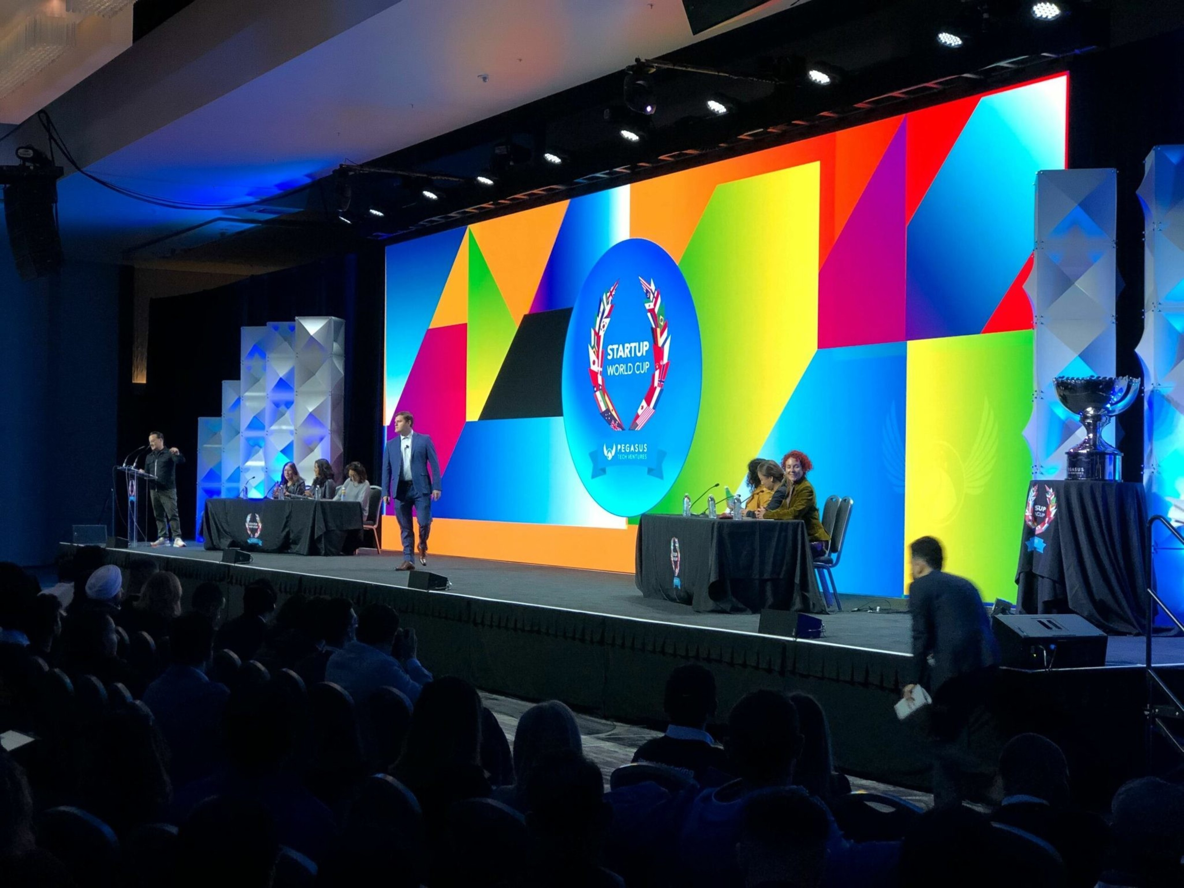 A colorful stage with a large "Startup World Cup" logo features judges seated at two tables, a speaker at a podium, and an audience in a dimly lit room.