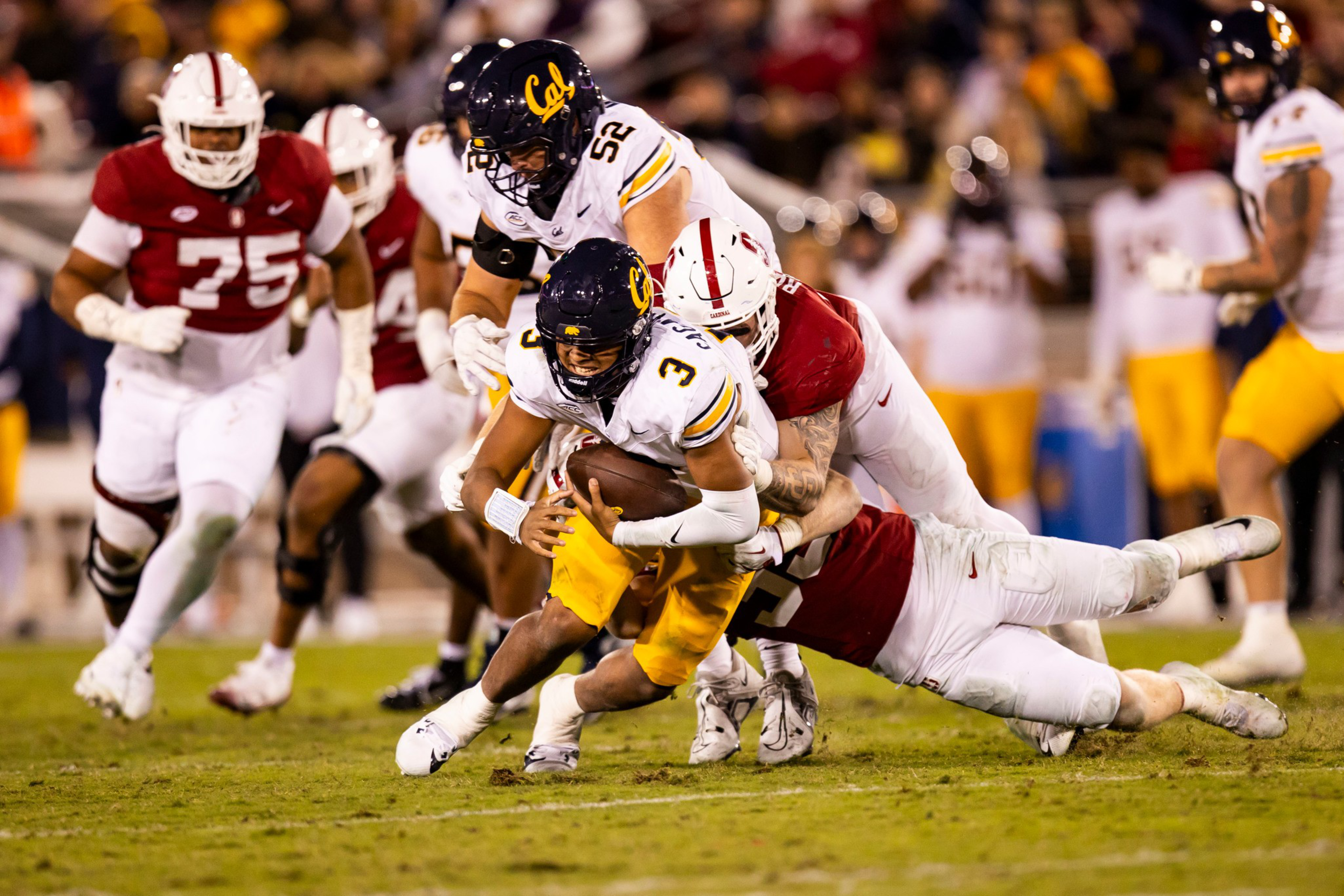 A football player in a white and yellow Cal uniform is being tackled by two players in red and white Stanford uniforms during a game.