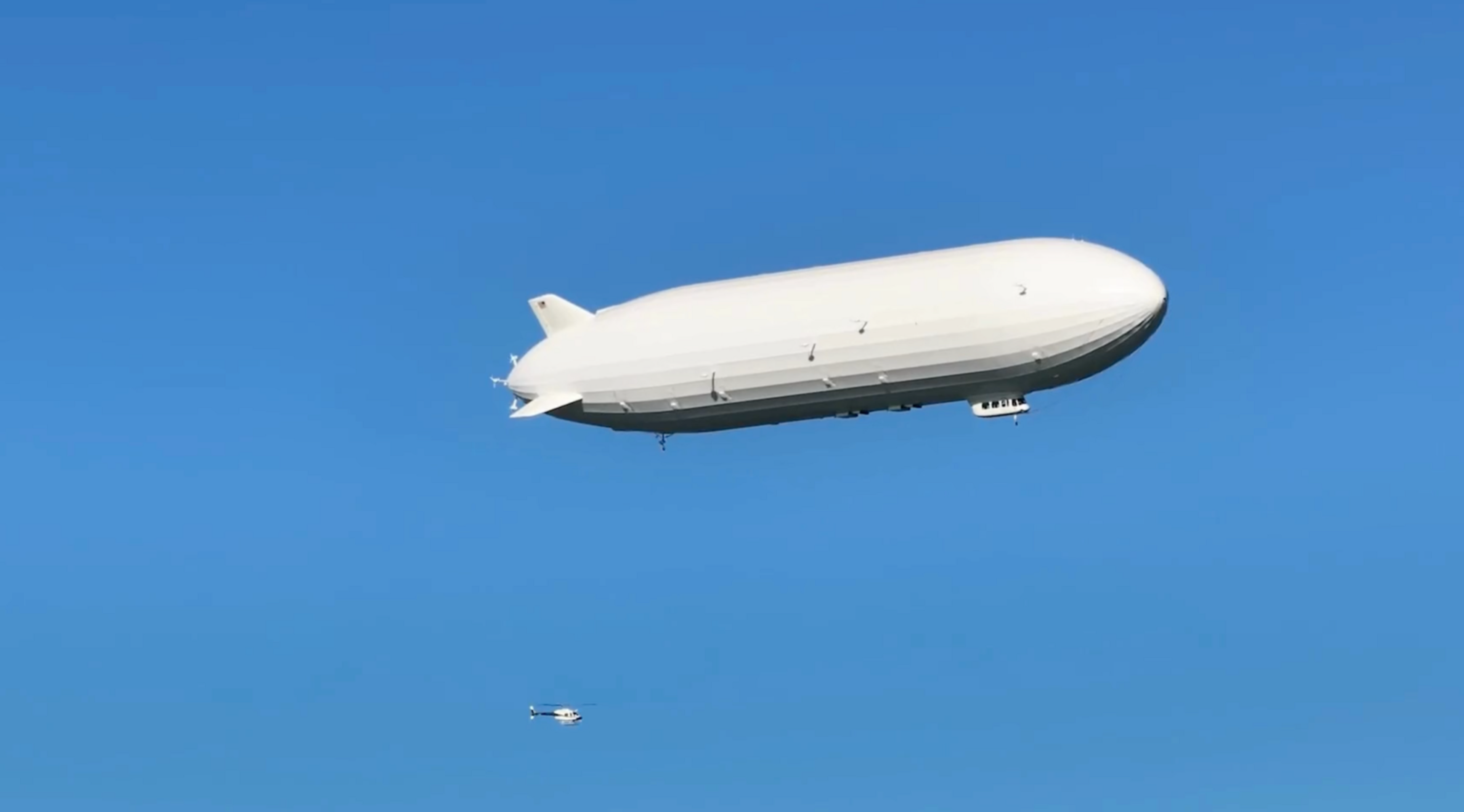 A large white blimp floats in a clear blue sky with a small helicopter visible below and beneath it.