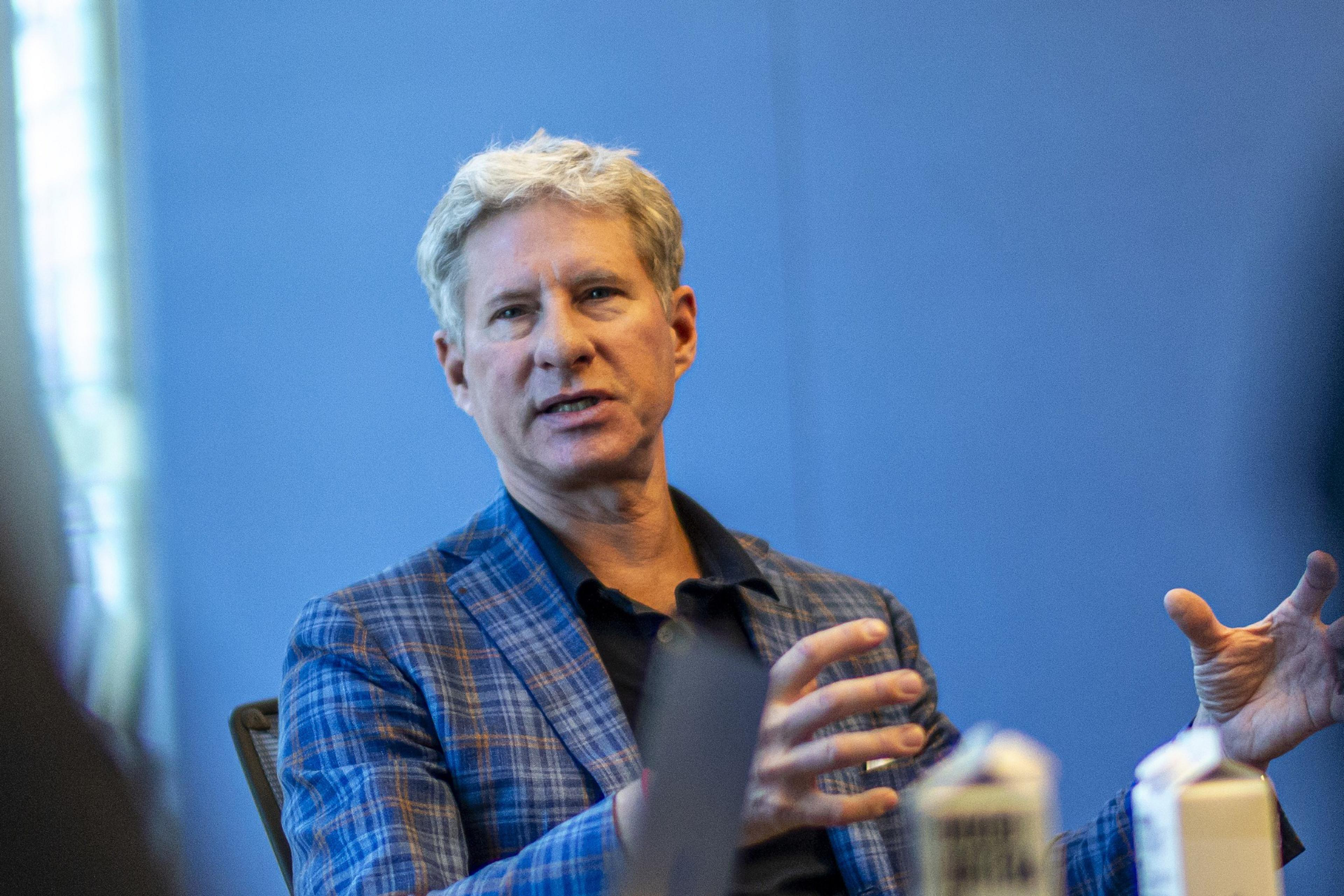 A man with gray hair wearing a blue plaid jacket gestures with his hands while speaking against a blue background.