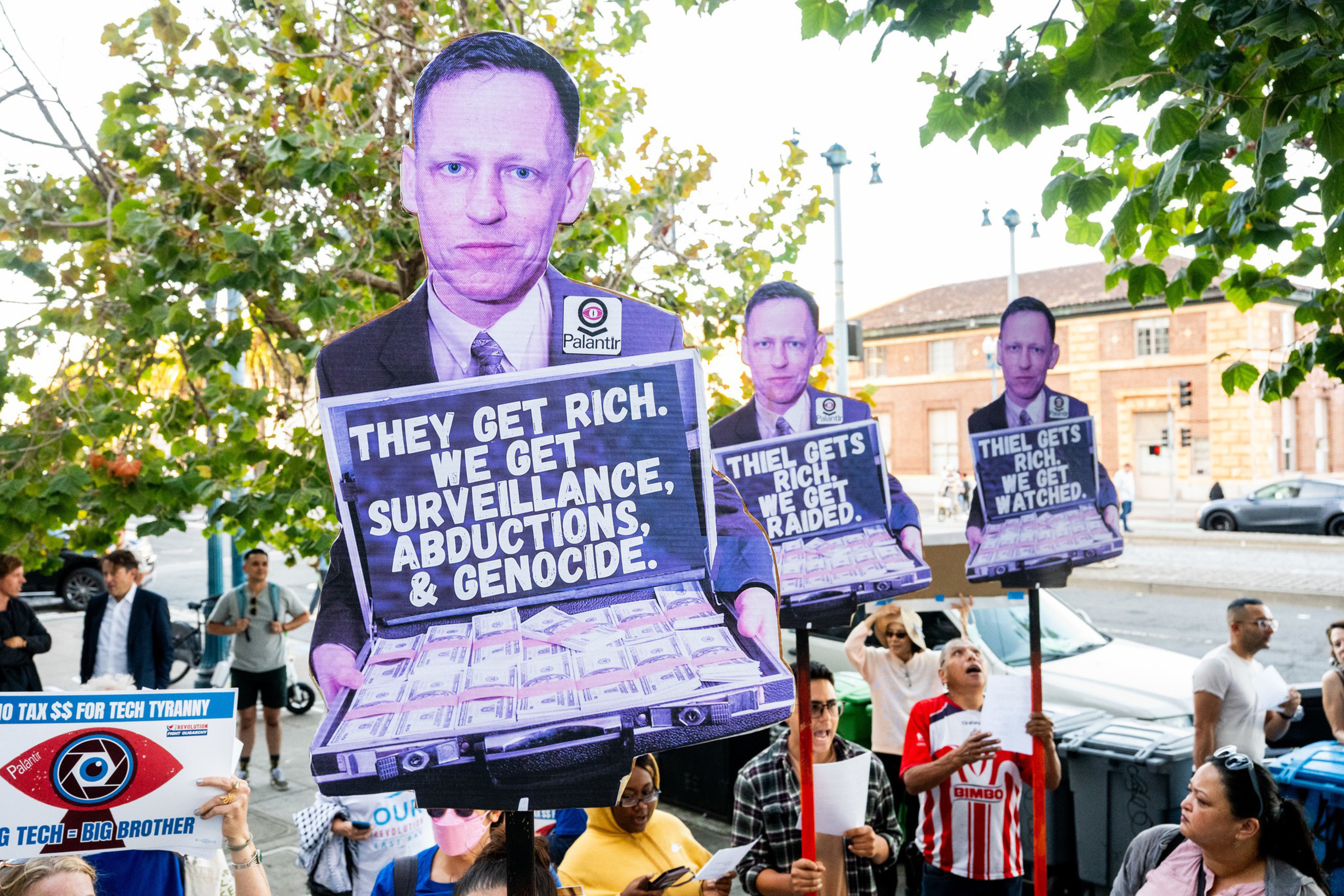 Three people stand outdoors holding protest signs criticizing surveillance, abductions, and genocide, with trees and buildings in the background.