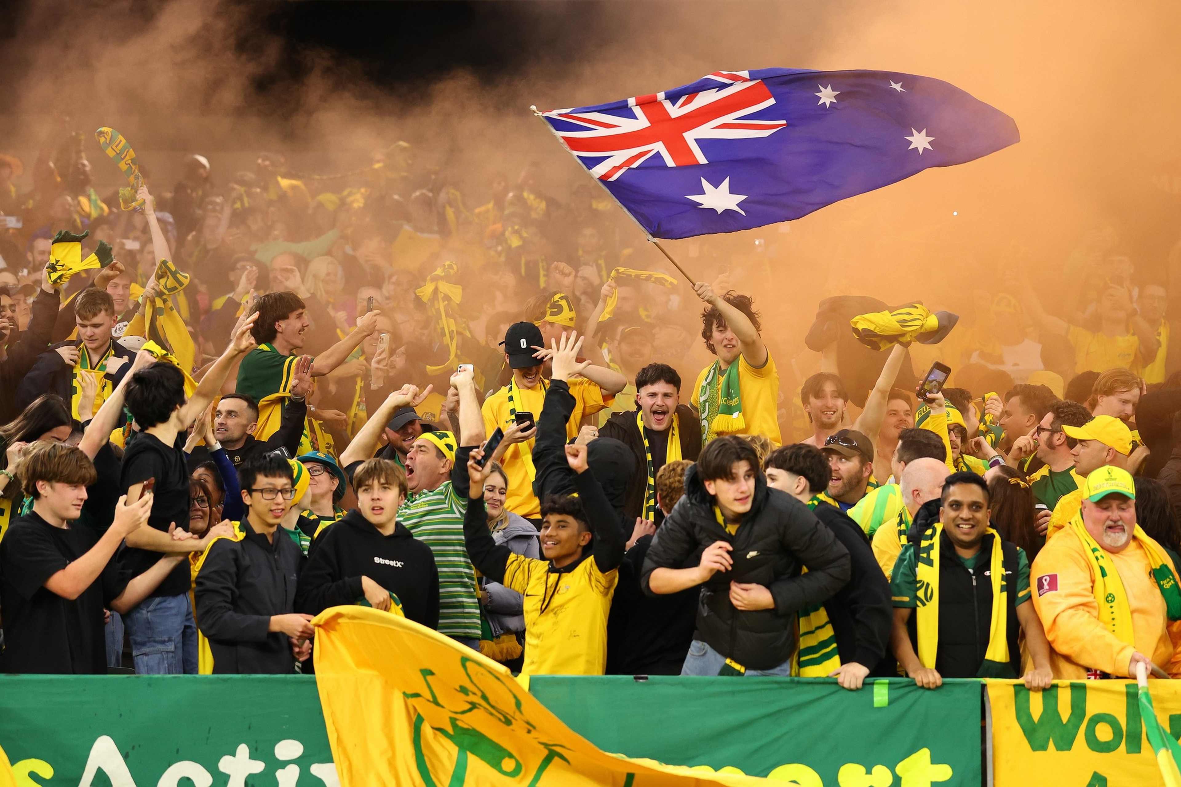 A lively crowd of Australian sports fans wave flags and wear yellow and ...