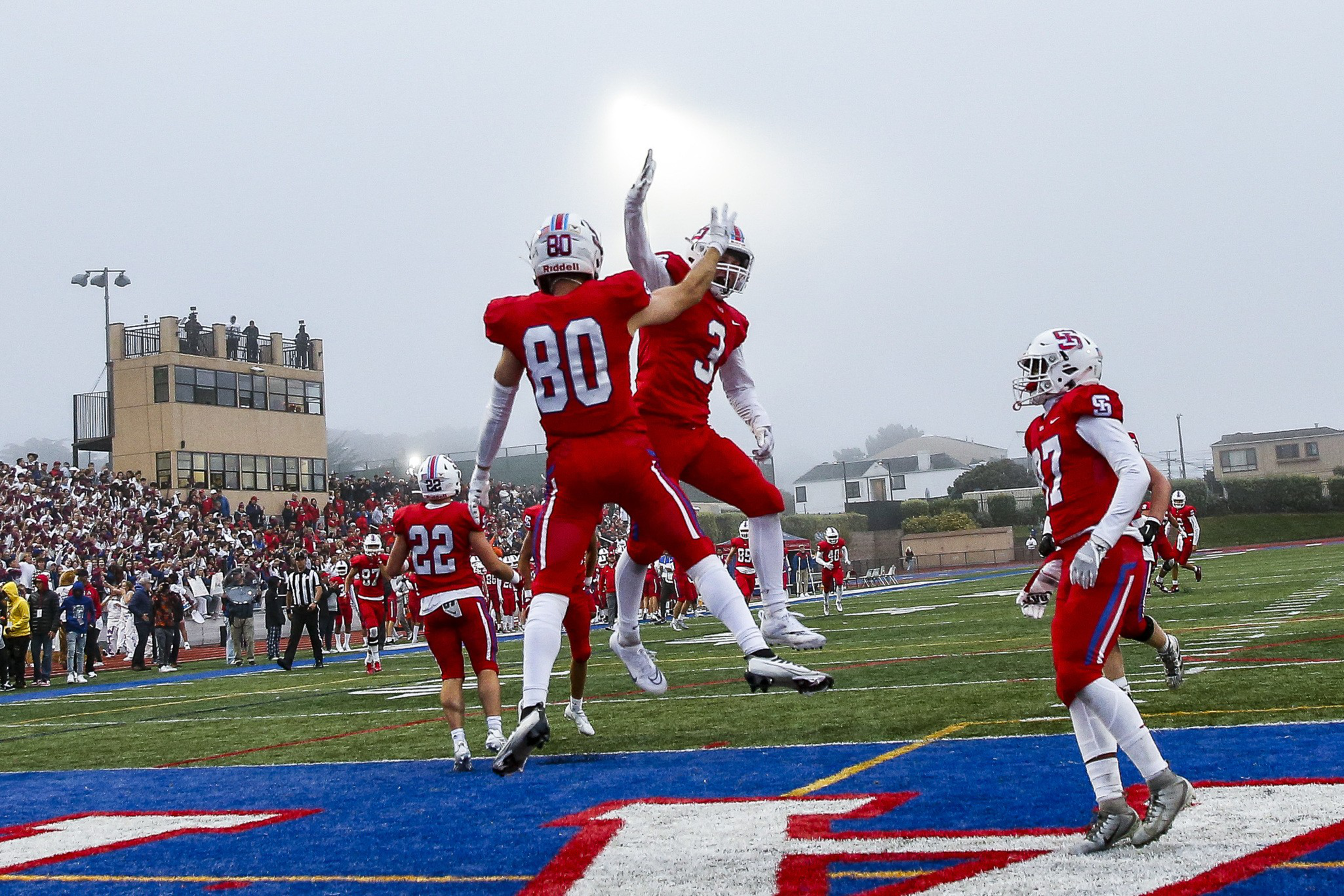 Two football players in red uniforms jump to high-five near the end zone, while teammates and a crowd watch in the foggy stadium background.