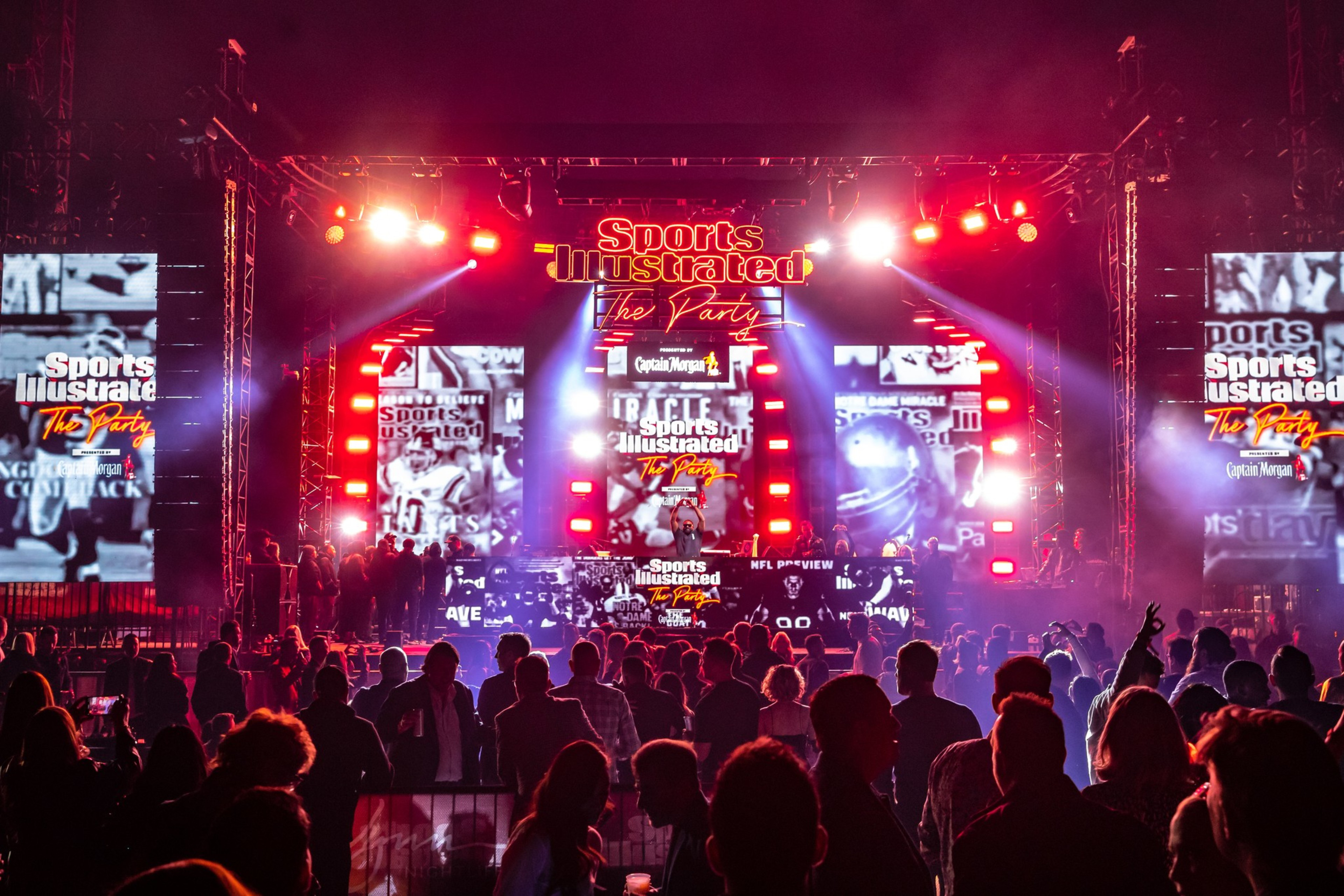 A crowded party with a large illuminated stage featuring “Sports Illustrated The Party” signs and bright red and white lights.