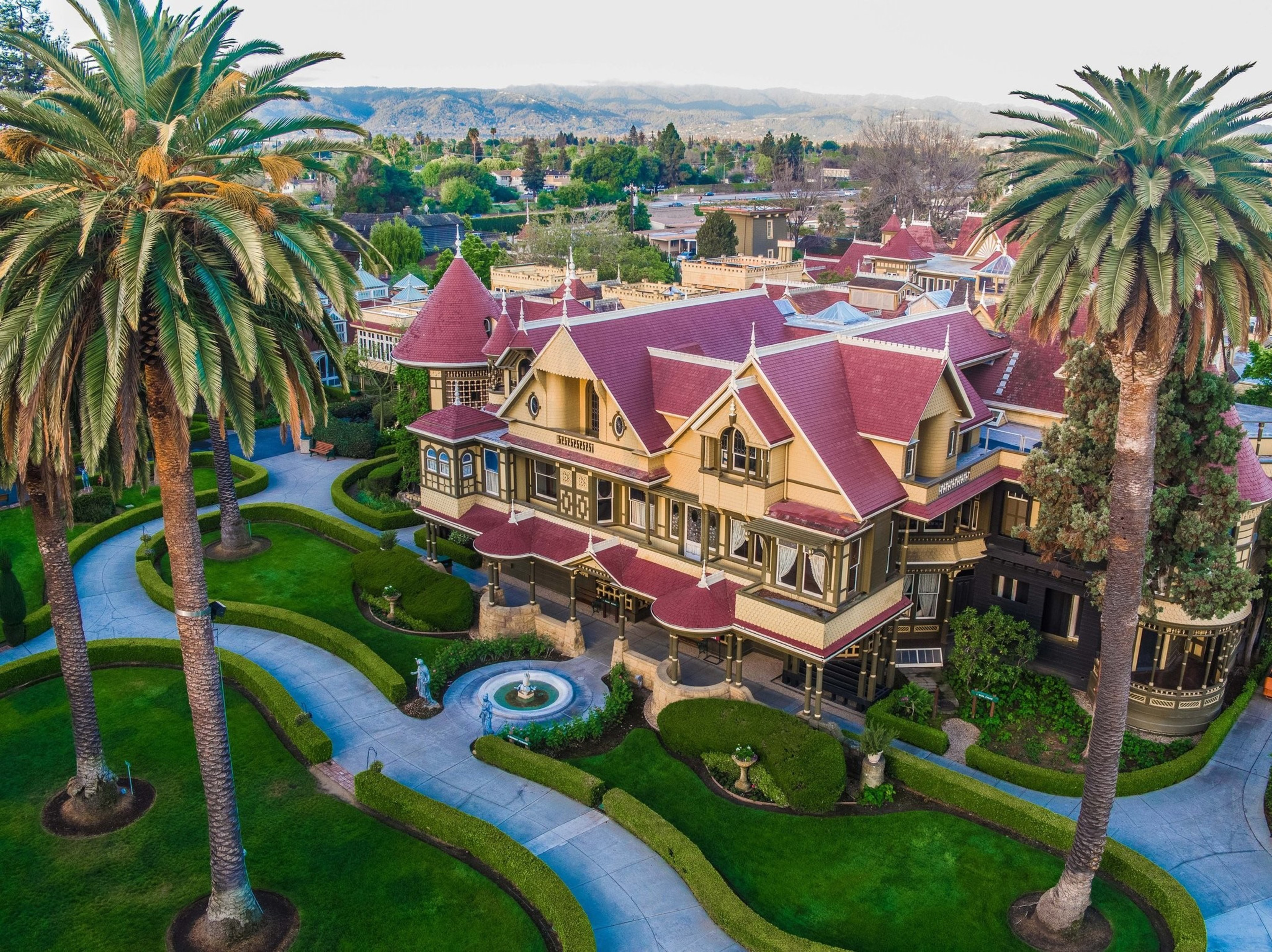 A large Victorian mansion with red roofs sits amidst manicured gardens, winding walkways, a central fountain, and tall palm trees on either side.