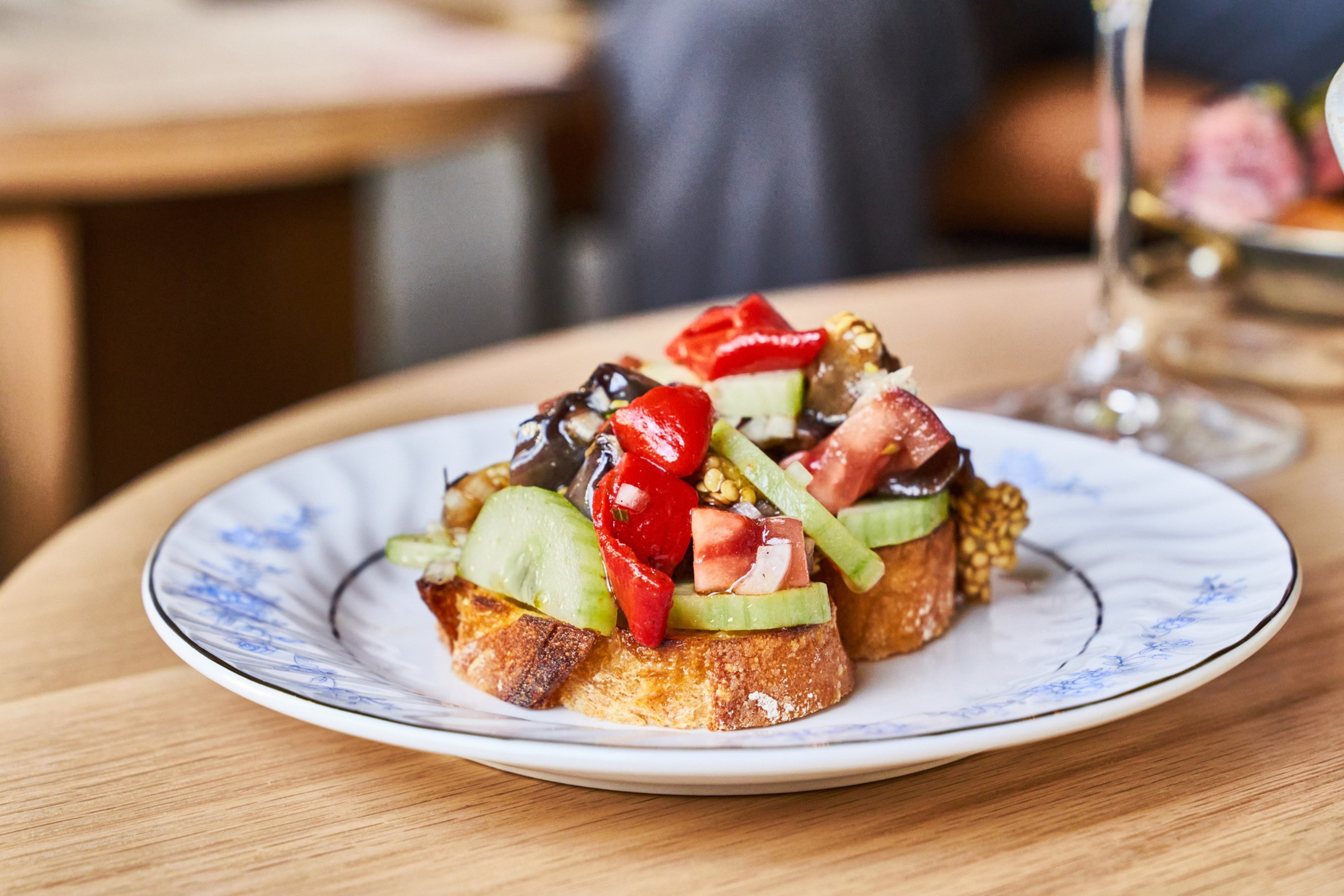 The image shows two slices of bread topped with cucumber, tomato, red peppers, and eggplant on a white plate with blue floral patterns.