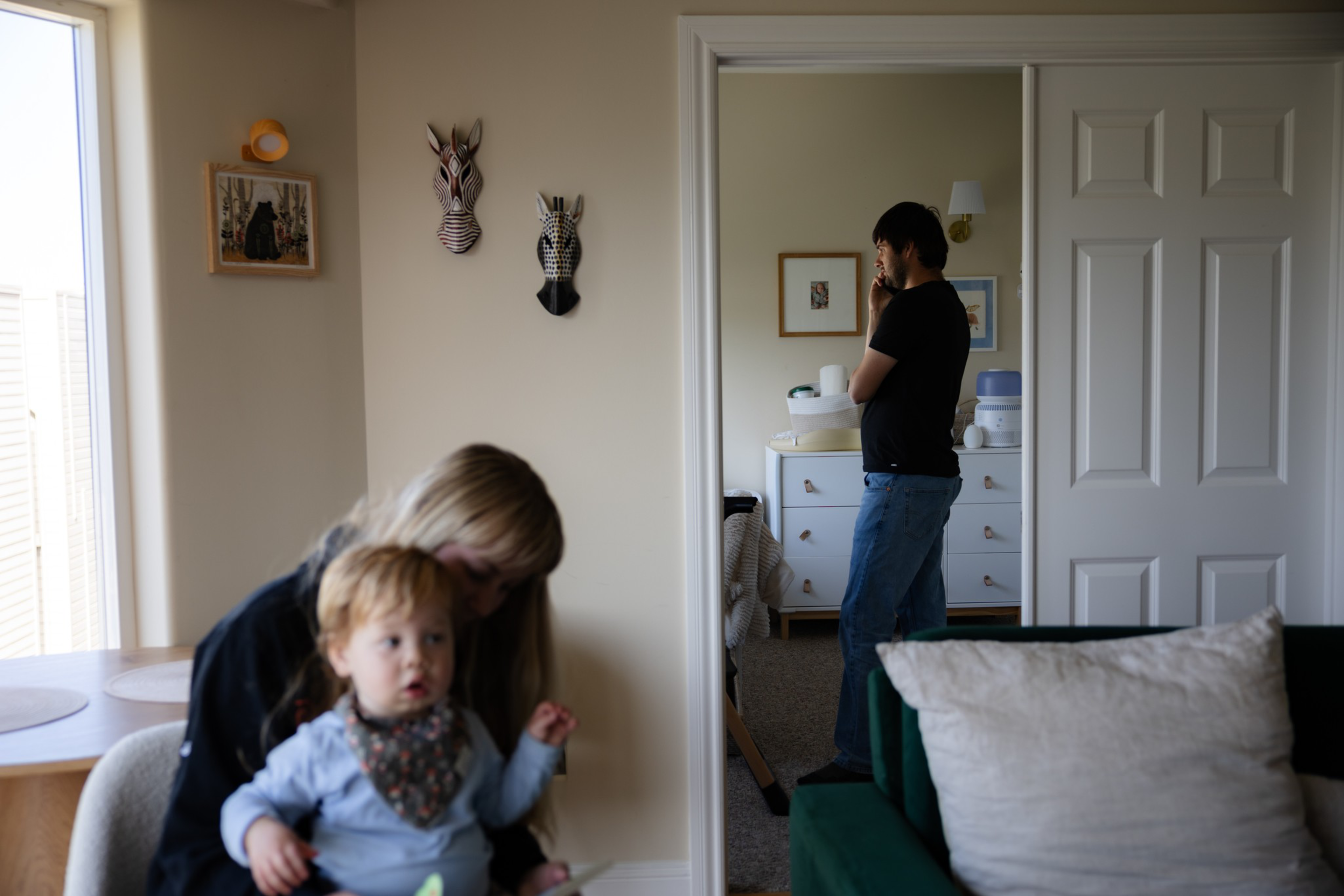 A woman holds a toddler near a wall with animal masks while a man stands in the next room on the phone; a green couch and a white pillow are visible.