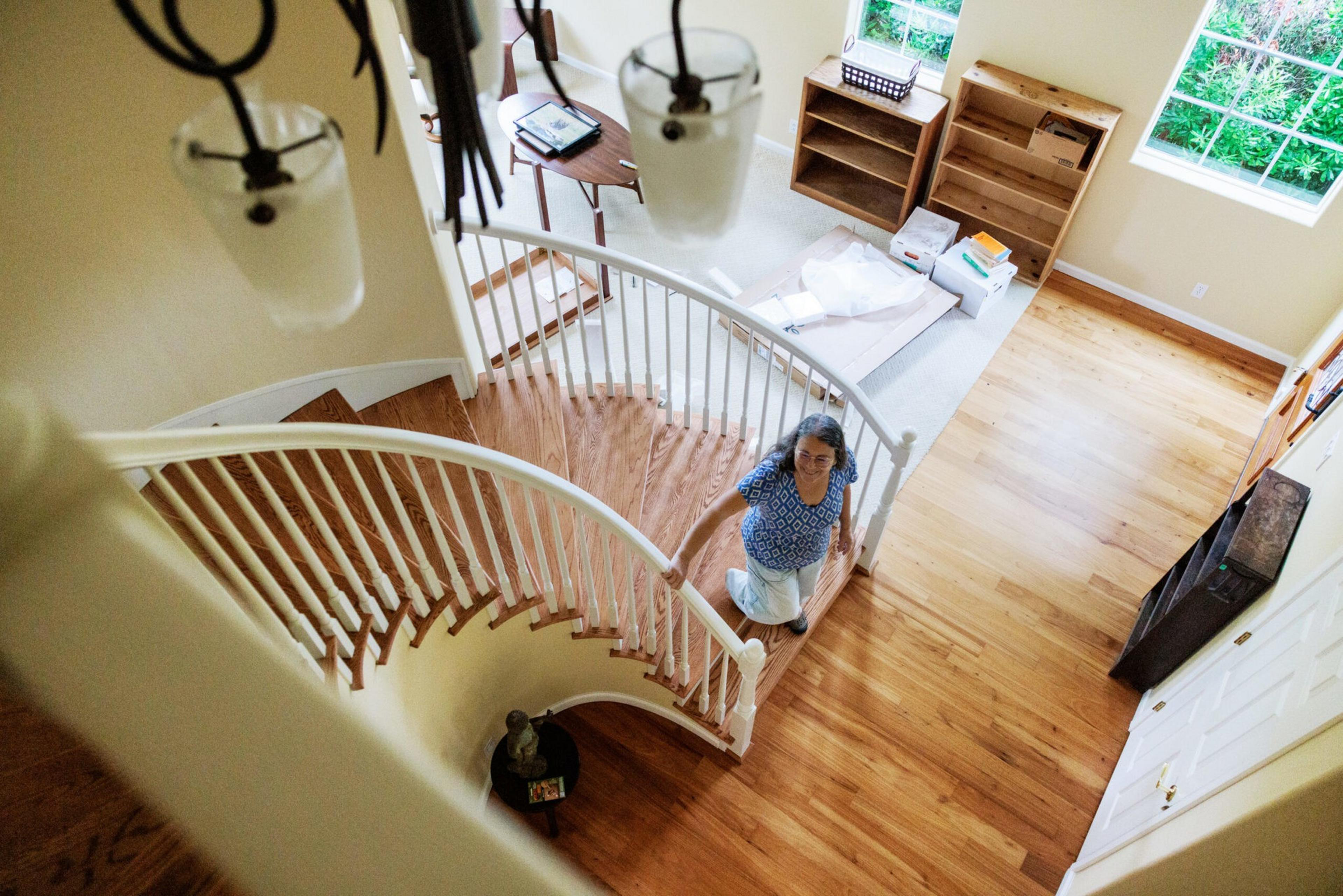 A woman with gray hair kneels on a curved wooden staircase railing inside a home with hardwood floors, natural light, and empty shelves.