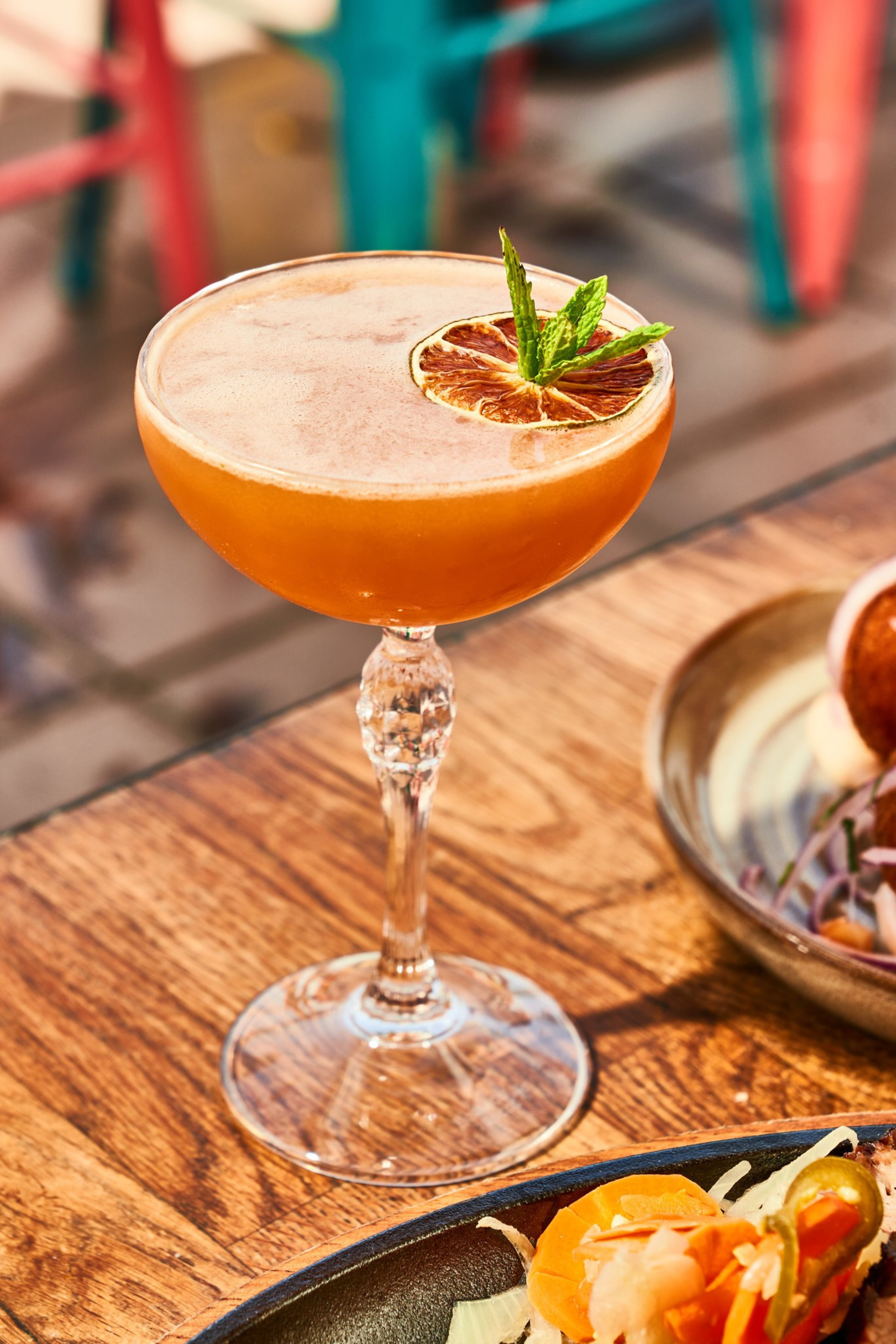 A cocktail in a decorative glass is garnished with a dried citrus slice and mint leaves, set on a wooden table next to a plate with sliced vegetables.