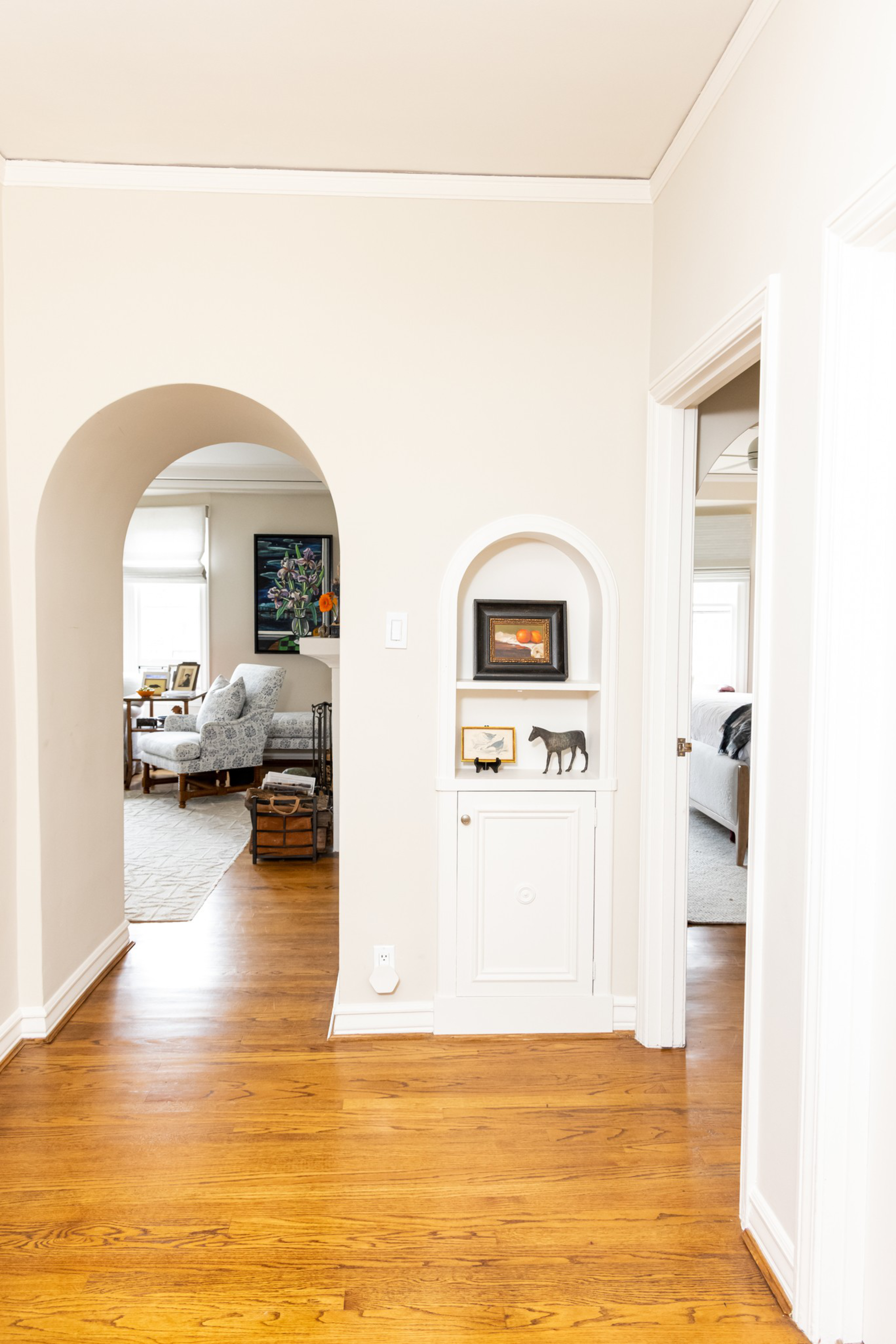 A hallway with light wooden floors leads to a living room through an archway and a bedroom on the right, with a built-in shelf displaying small framed art and figurines.
