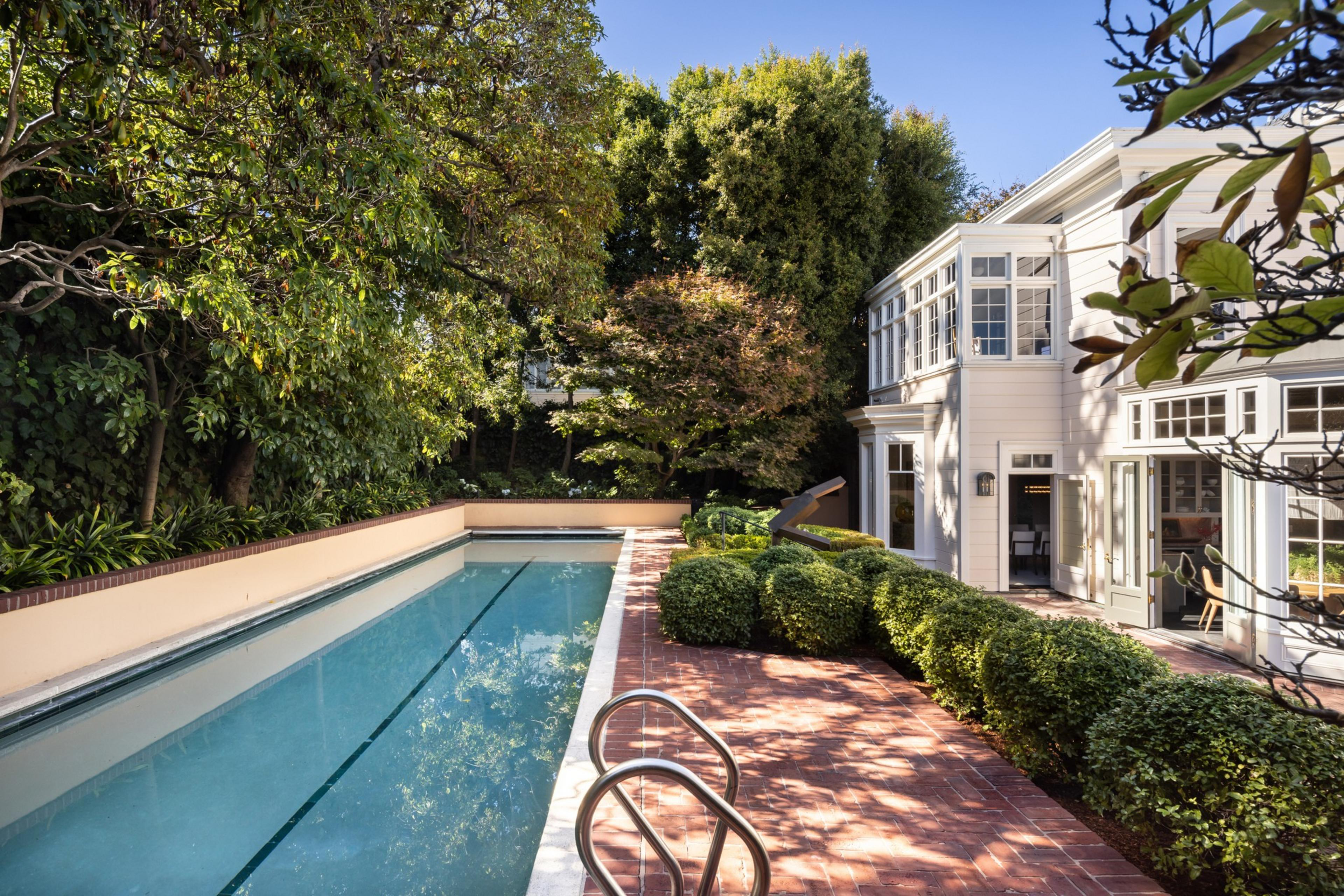 A long, narrow swimming pool is surrounded by a brick patio, lush greenery, and a modern white house with large windows under a clear blue sky.