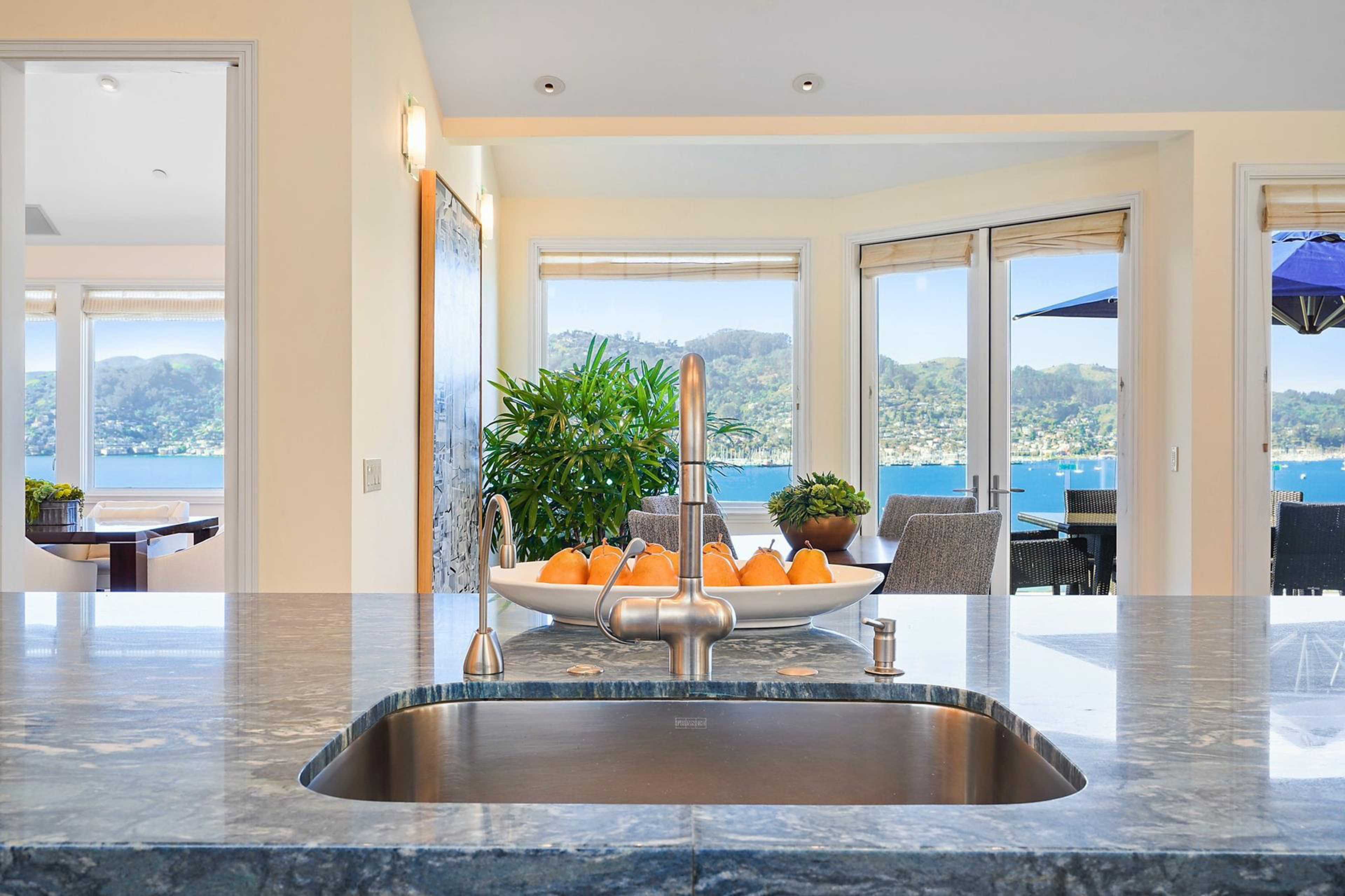 A kitchen sink with a stainless steel faucet sits on a marble countertop, overlooking a dining area, plants, and large windows with a view of water and hills.