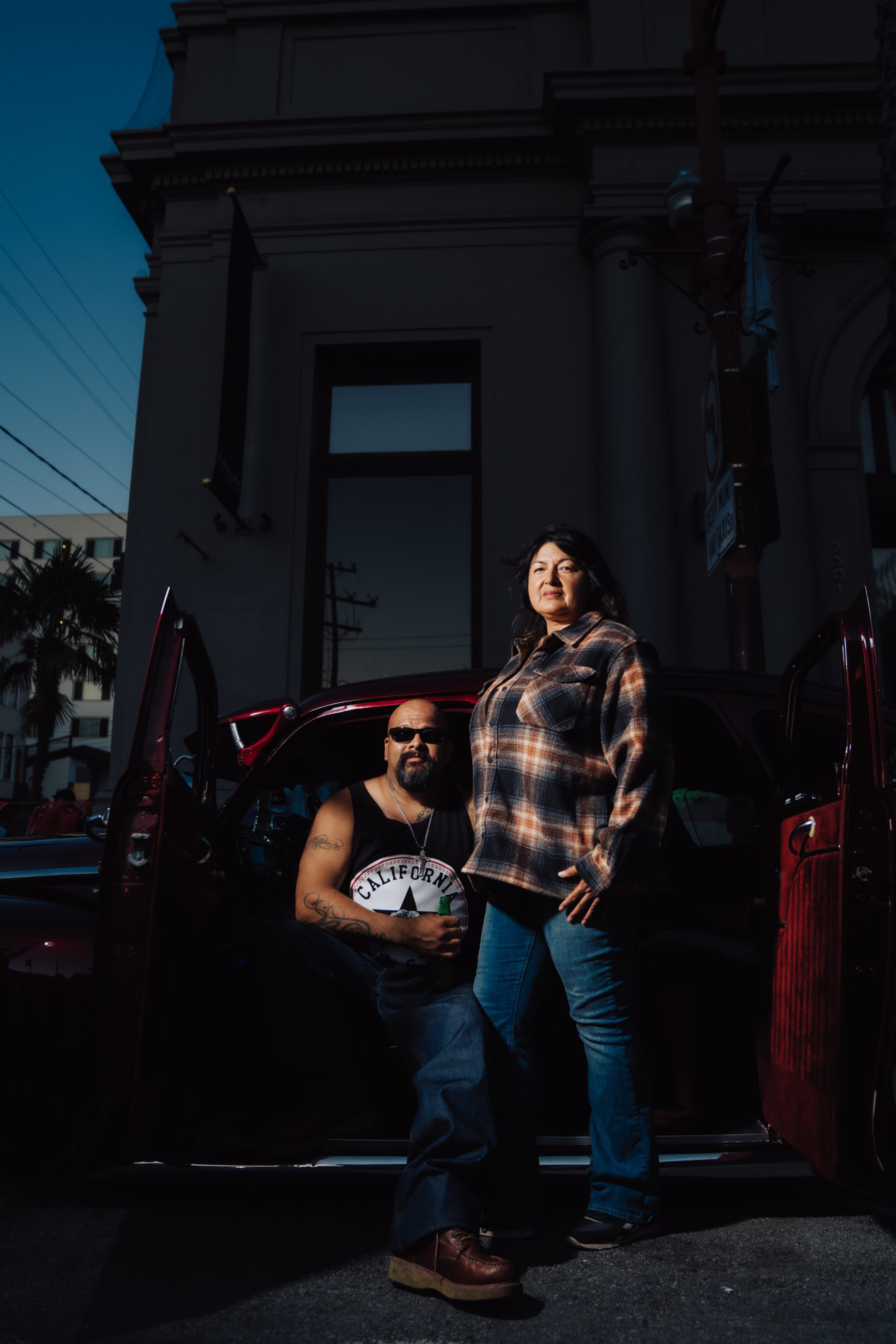A man with sunglasses and a sleeveless shirt sits in a vintage car, while a woman in a plaid shirt and jeans stands beside him in front of a building.