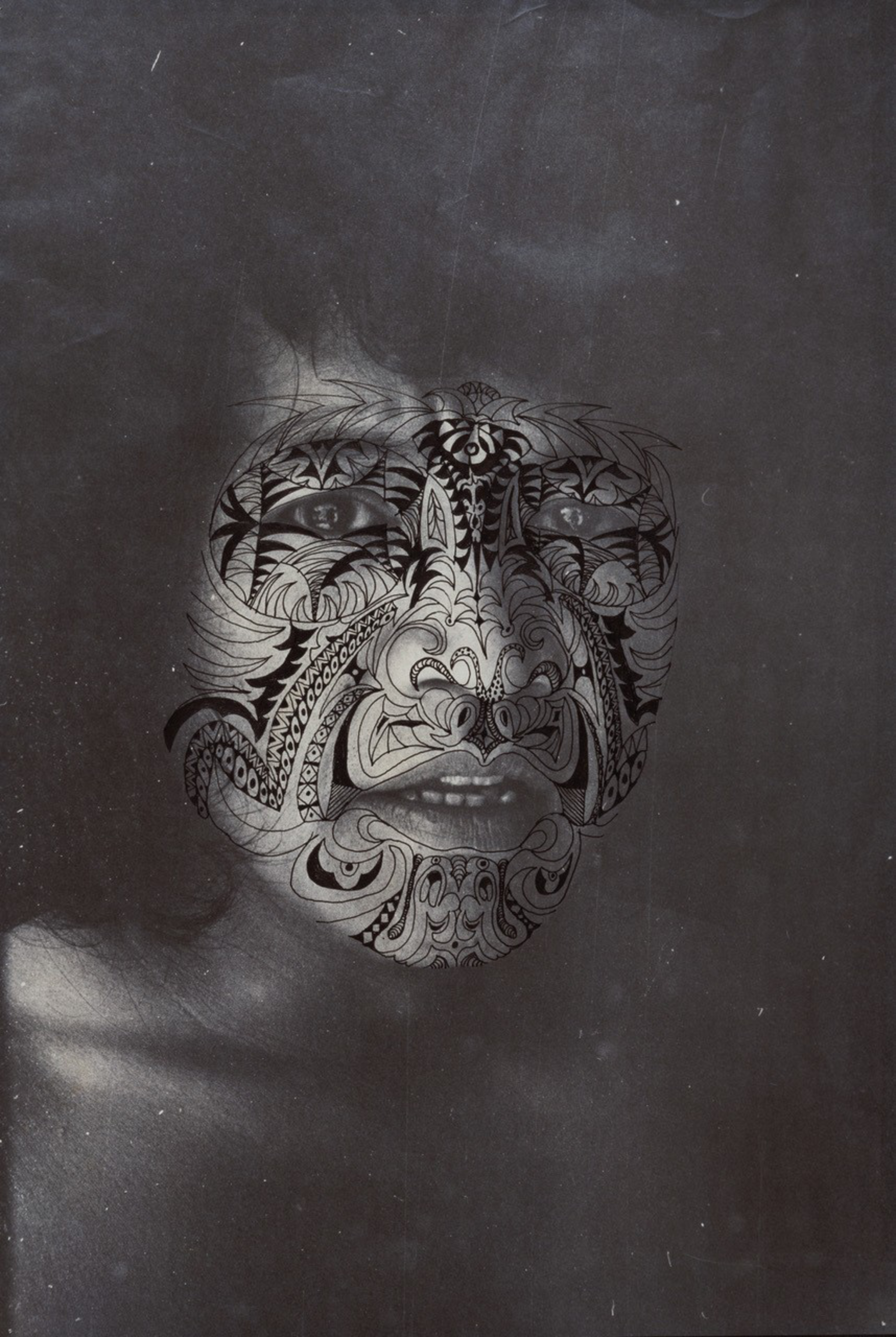 A face with intricate, dark tribal tattoo patterns covering it, blending with the grayscale tones of the photograph, creating a mysterious, artistic effect.